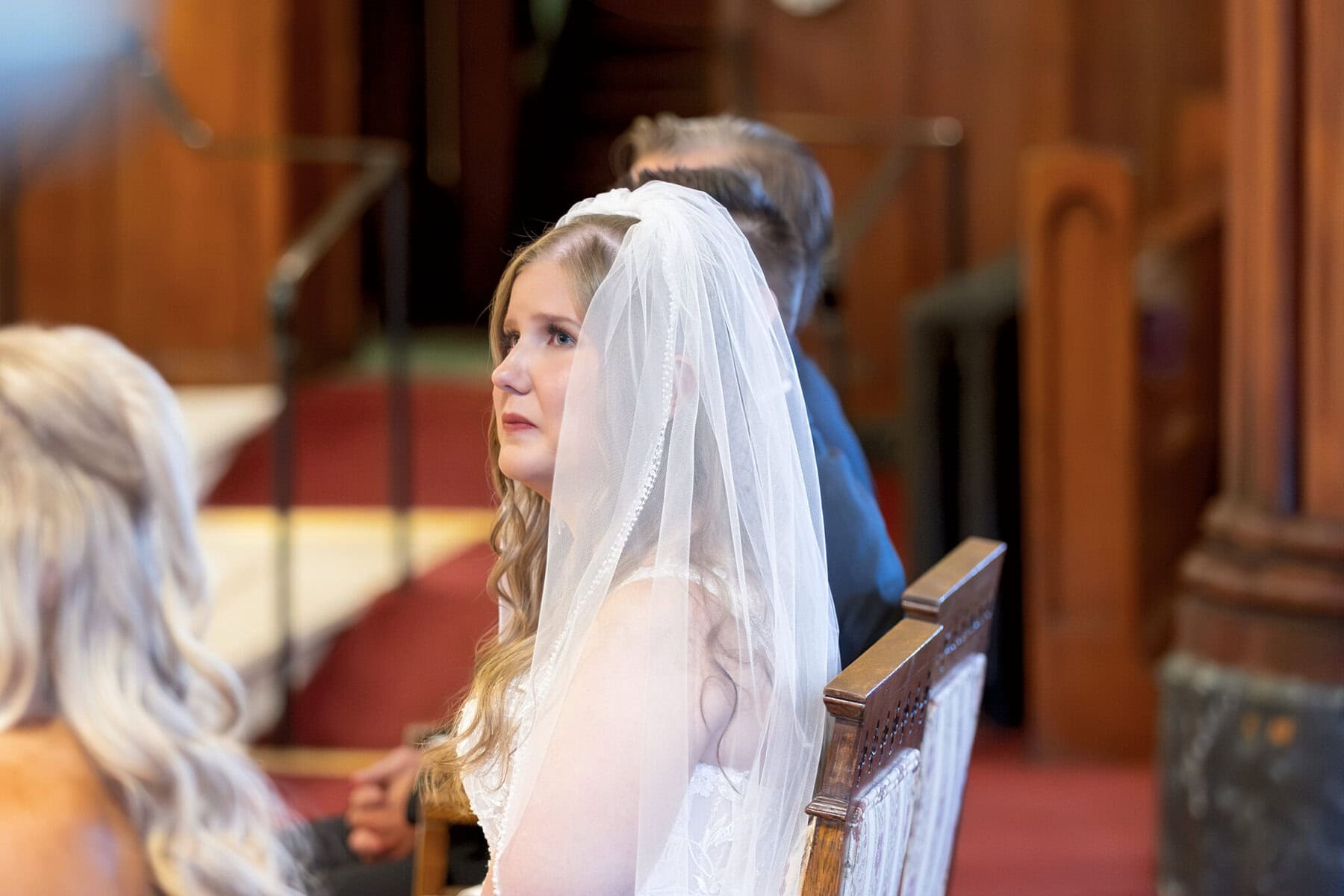 Wedding At St. Mary's Church in North Attleborough 32 A bride in a white dress and veil sits indoors at St. Mary's church, looking forward, with guests seated nearby on wooden chairs.