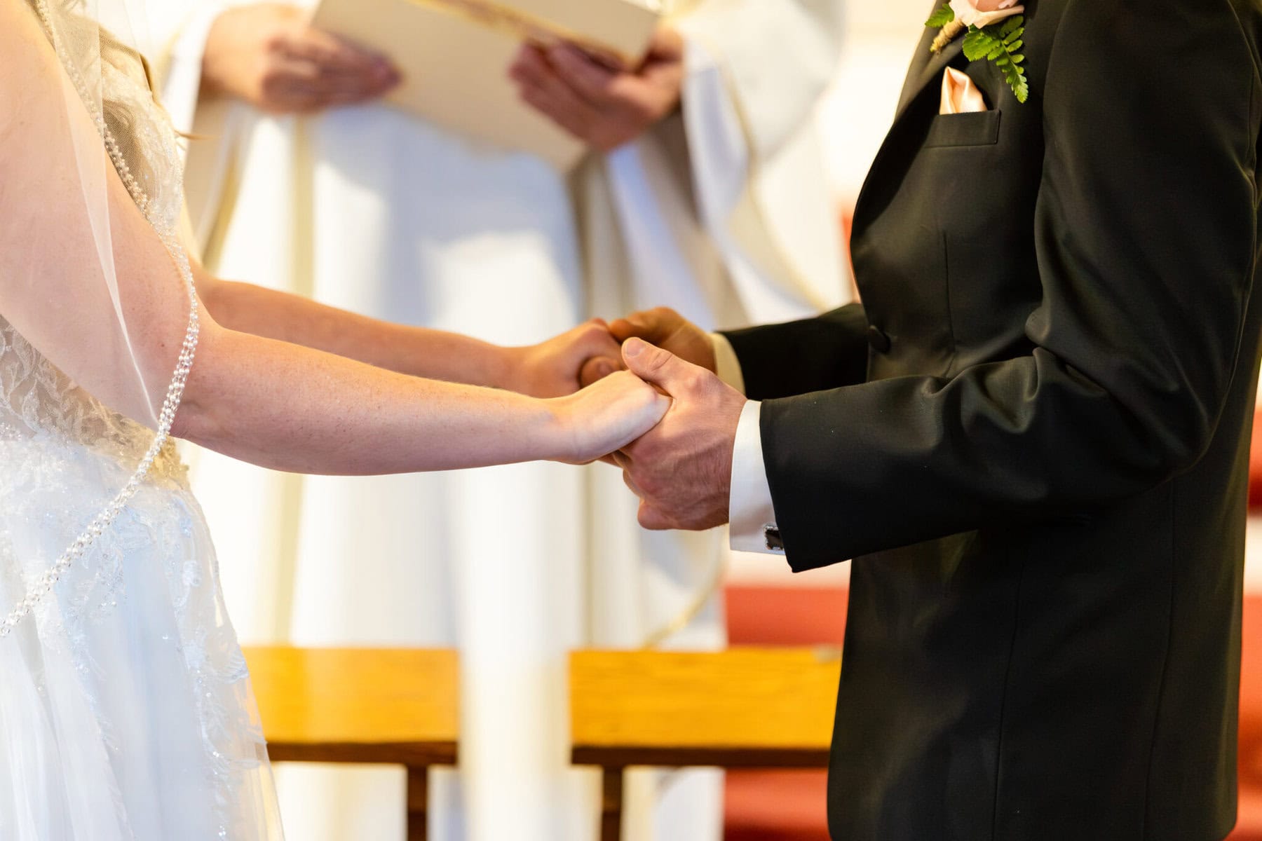 Wedding At St. Mary's Church in North Attleborough 35 A bride and groom hold hands during a wedding ceremony at St. Mary's church, with an officiant standing in the background holding papers.