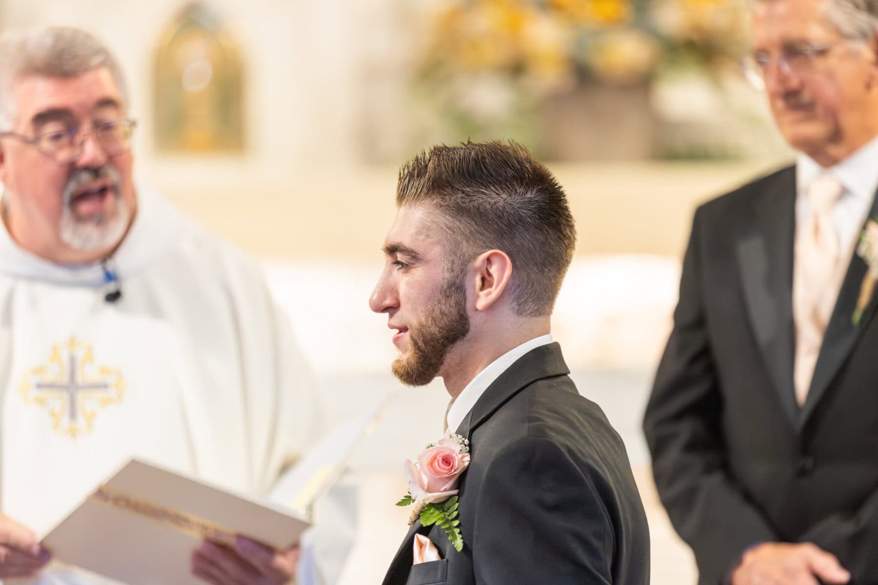 Wedding At St. Mary's Church in North Attleborough 36 A groom in a black suit with a pink rose boutonniere stands at his North Attleborough wedding in St. Mary's church beside a priest and another man.