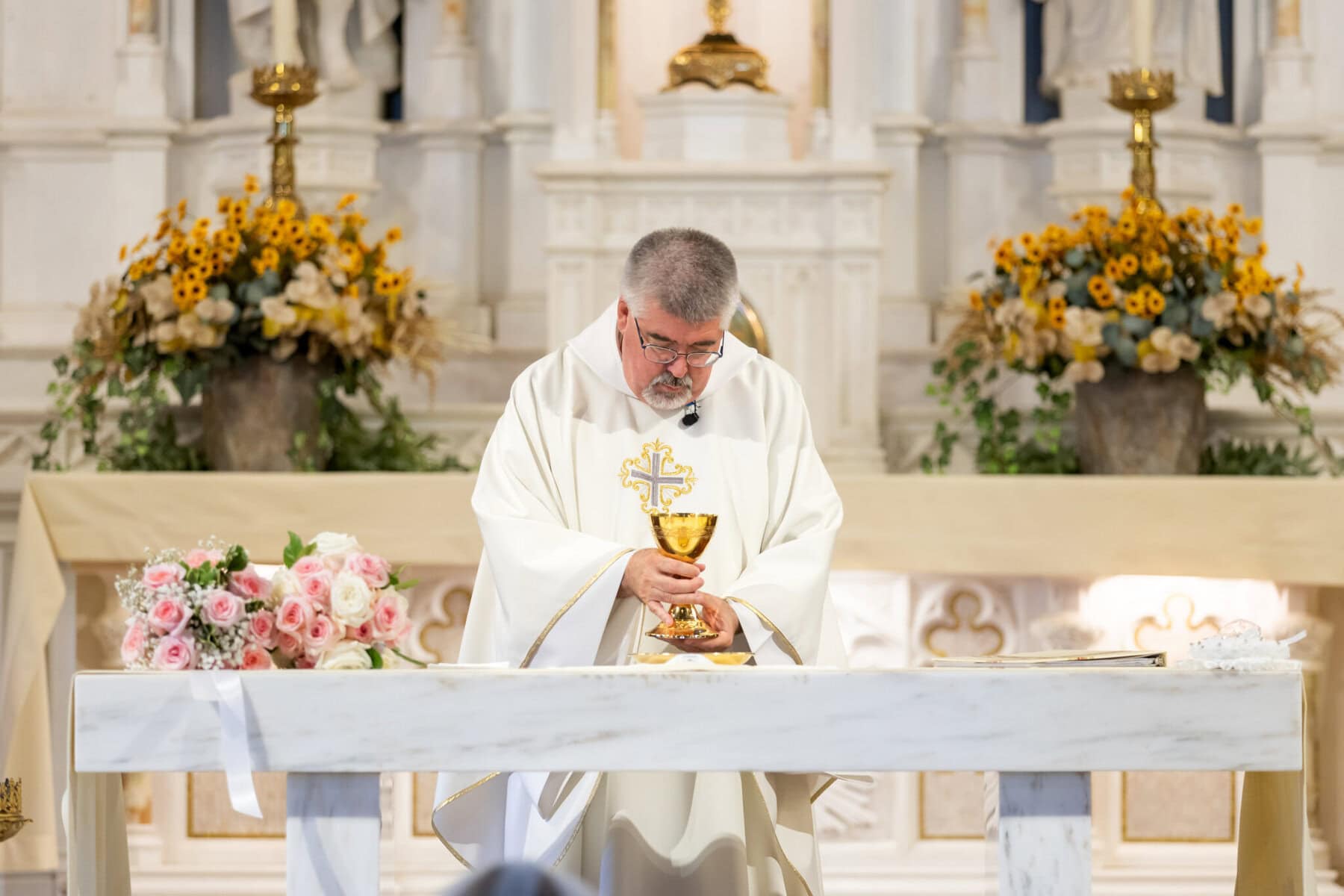 Wedding At St. Mary's Church in North Attleborough 40 A priest in white robes holds a chalice at a flower-adorned altar during a wedding at St. Mary's Church in North Attleborough.