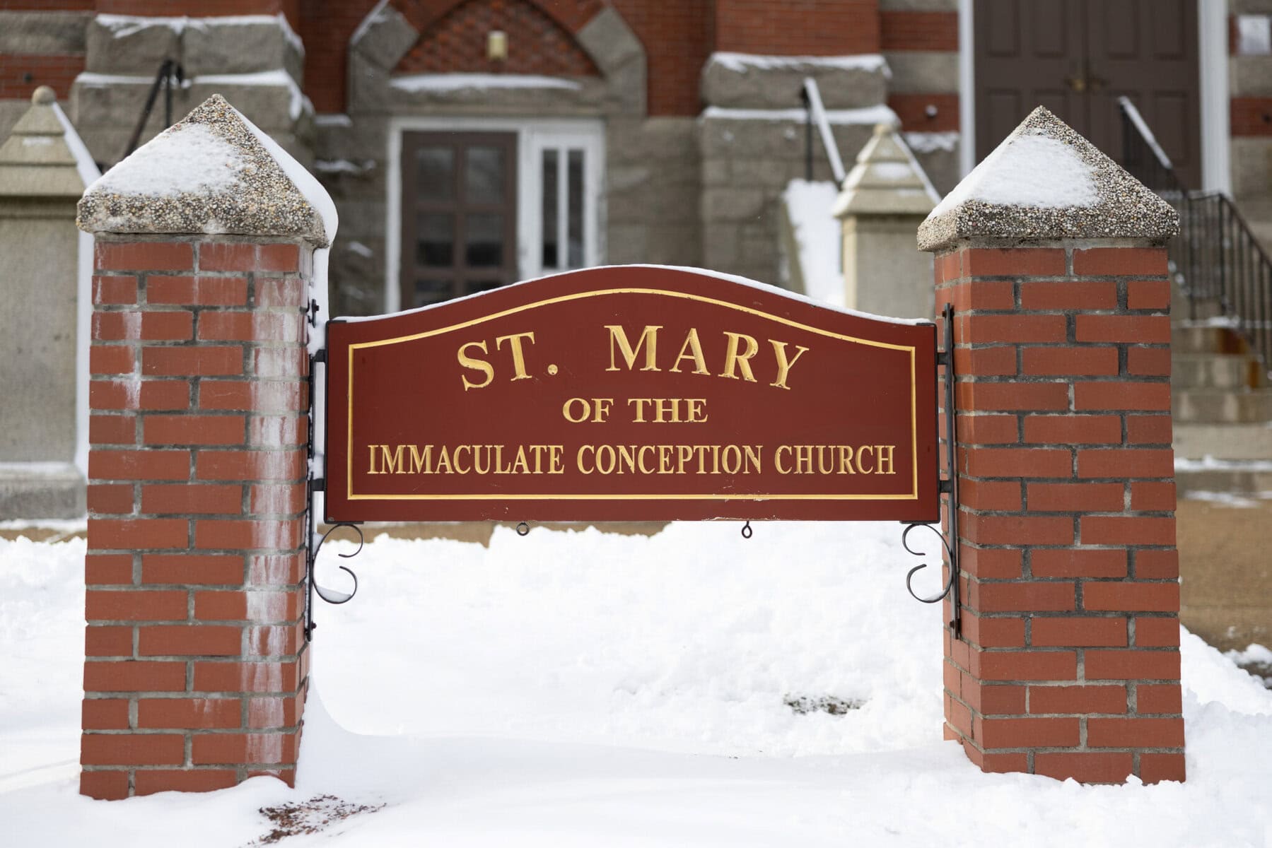 Wedding At the Transfiguration of the Lord Parish 3 A red and gold sign reading “St. Mary of the Immaculate Conception Church” stands between brick pillars, a serene spot for a Wedding amid snow.