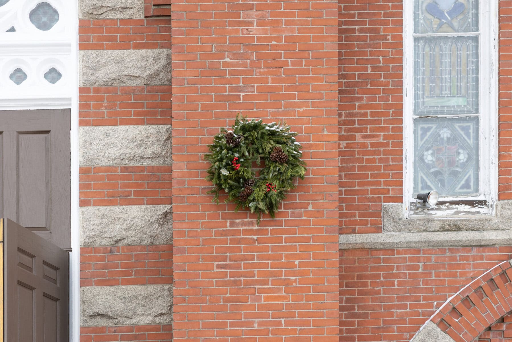 Wedding At the Transfiguration of the Lord Parish 5 A green holiday wreath with pinecones and red berries hangs on a red brick wall near a stained glass window at Transfiguration of the Lord.