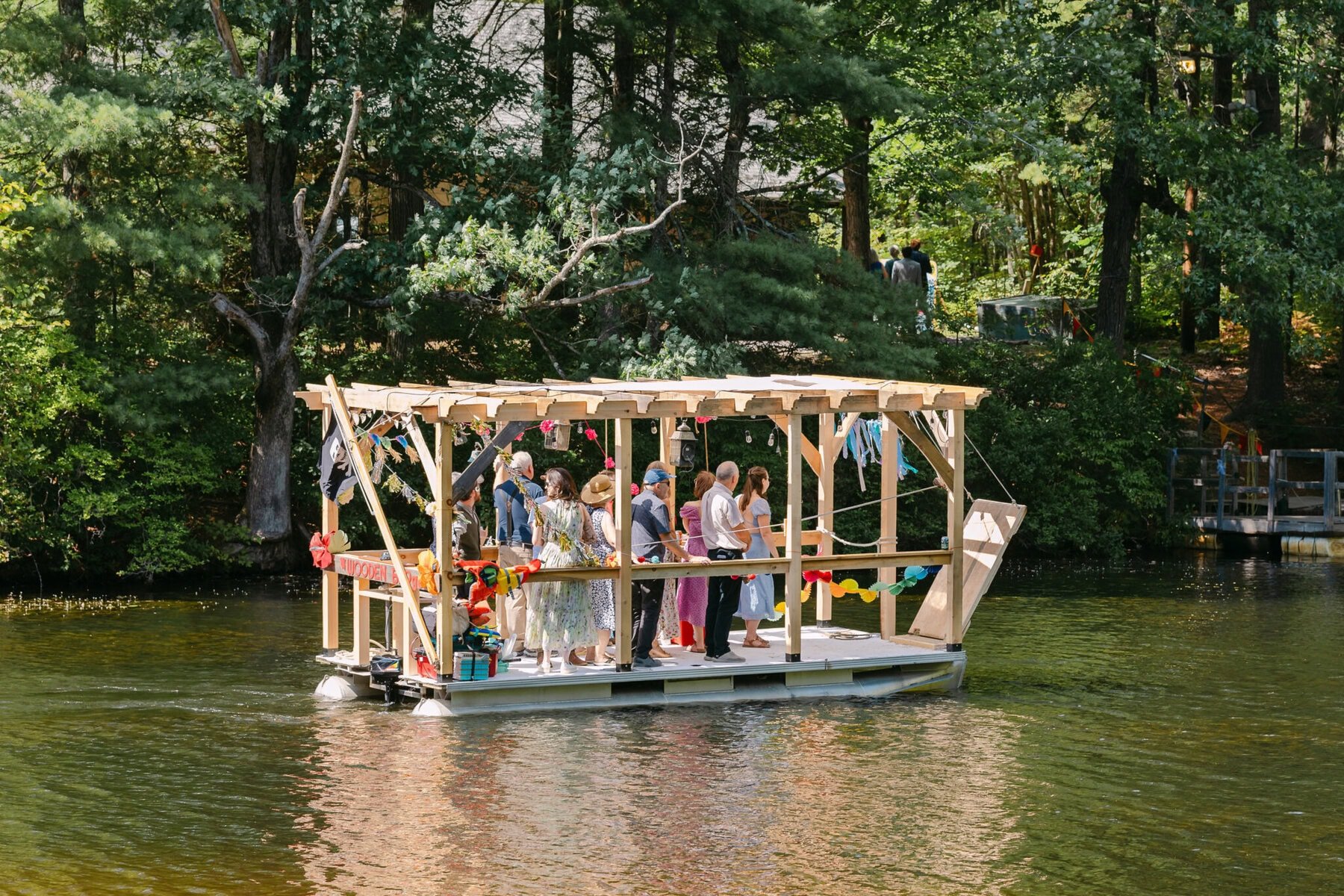 A Worcester Wedding at Cook Pond 6 A group of people stand on a wooden raft with colorful bunting, floating on Cook Pond’s calm river, surrounded by trees.