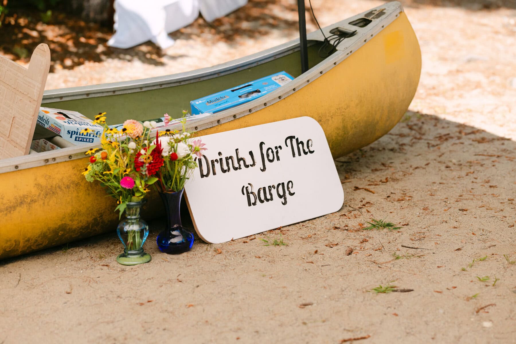 A Worcester Wedding at Cook Pond 2 A yellow canoe on sand holds drink boxes at a Worcester Wedding by Cook Pond. A sign reads "Drinks for the Barge." Flowers sit nearby.