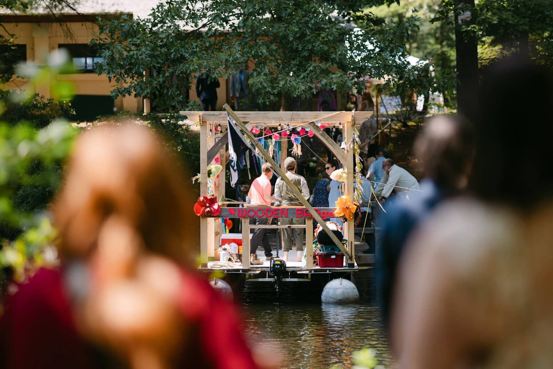 A Worcester Wedding at Cook Pond 7 Guests gather on a small wooden raft decorated with items, floating on Cook Pond at a Worcester Wedding, with blurred figures in front.