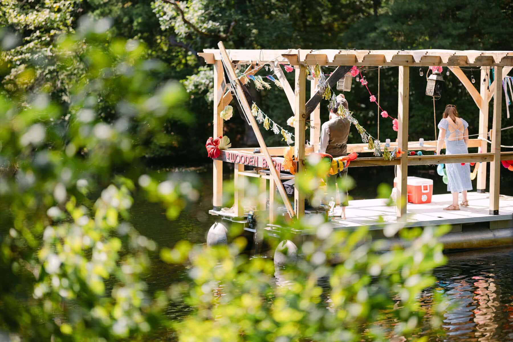 A Worcester Wedding at Cook Pond 8 Two people stand on a decorated wooden raft floating on Cook Pond, surrounded by green trees—a picturesque Worcester Wedding scene.