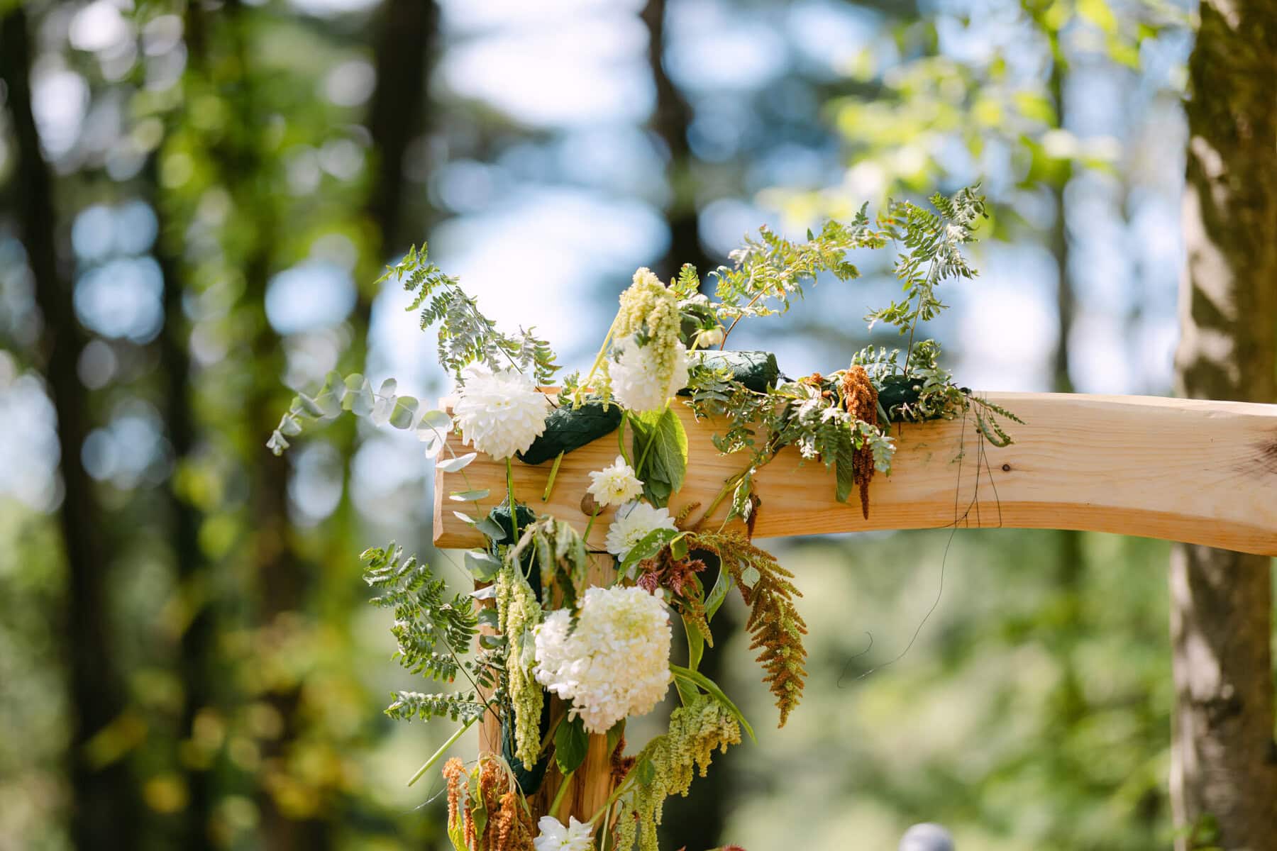 A Worcester Wedding at Cook Pond 13 A wooden arch adorned with white flowers and foliage stands at a Cook Pond Worcester Wedding, trees softly blurred in the background.