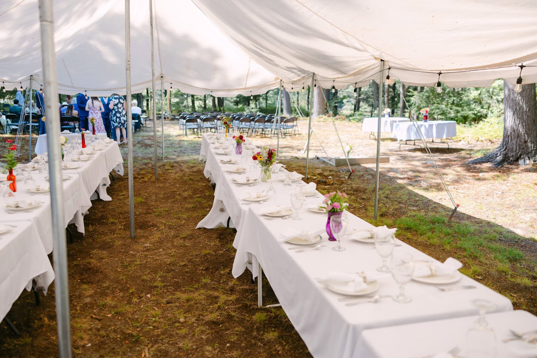 A Worcester Wedding at Cook Pond 16 Two long banquet tables with white linens await a charming Worcester Wedding at Cook Pond, set beneath a grand tent among trees.