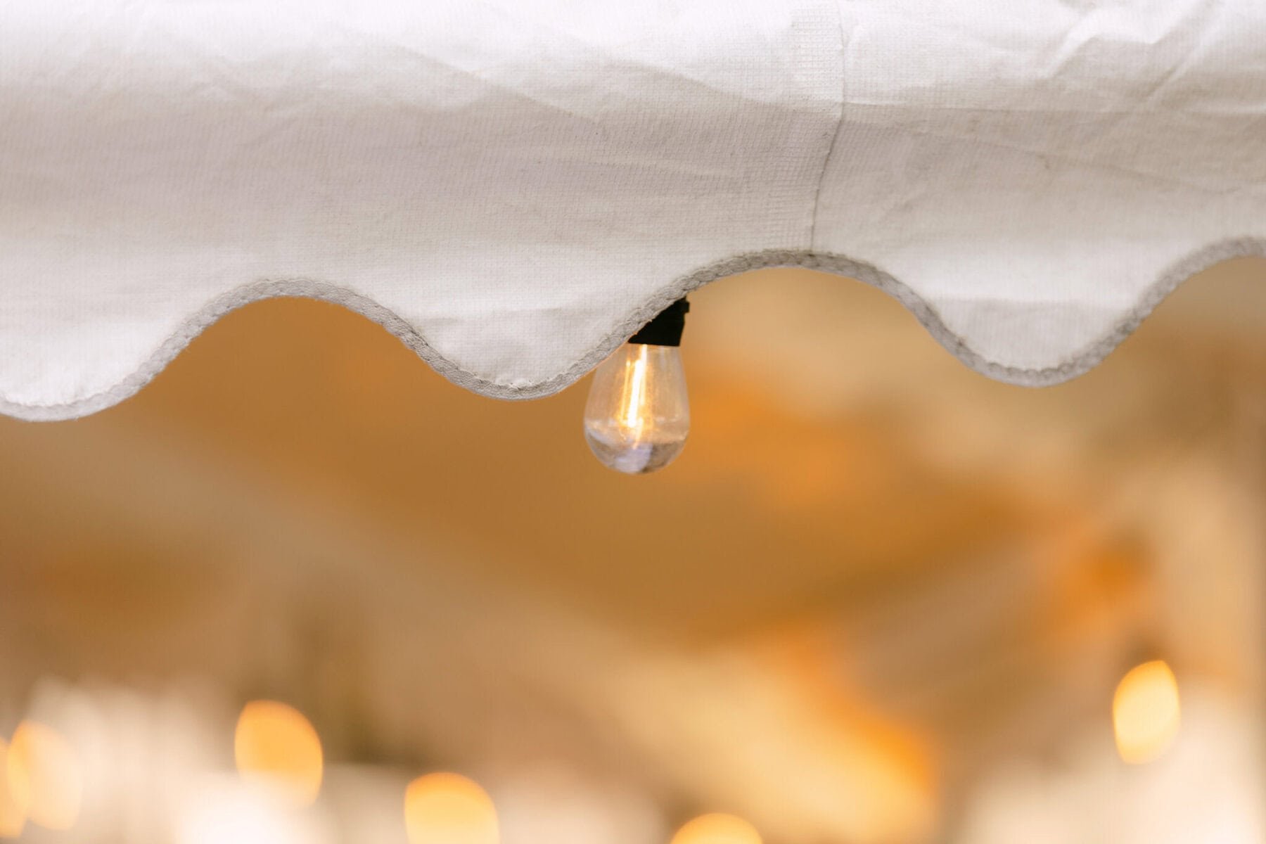 A Worcester Wedding at Cook Pond 20 A clear light bulb hangs from the scalloped edge of a white canopy at a charming Worcester wedding, with warm blurred lights in the background.