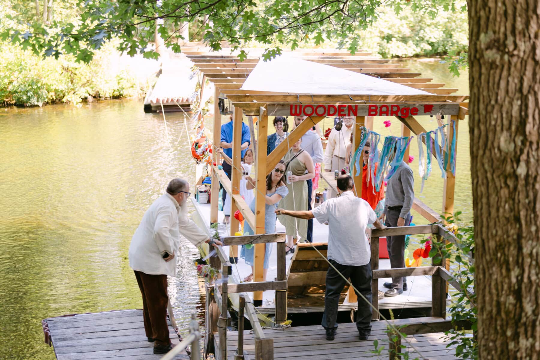 A Worcester Wedding at Cook Pond 10 A group stands on a dock by Cook Pond, near a decorated wooden barge, surrounded by trees and greenery.