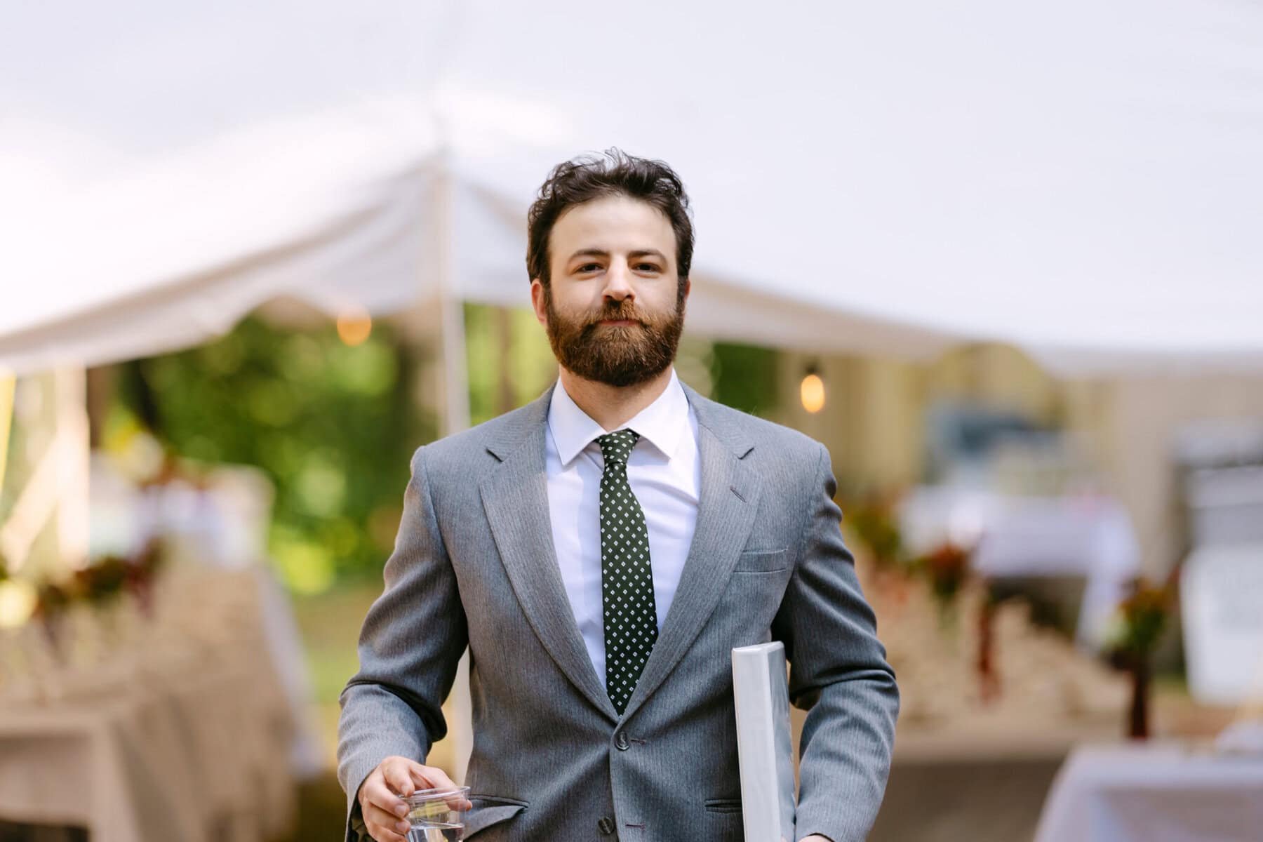 A Worcester Wedding at Cook Pond 23 A man in a gray suit and polka dot tie holds a laptop and glass, walking outdoors at a Worcester Wedding near tables set under a white tent.