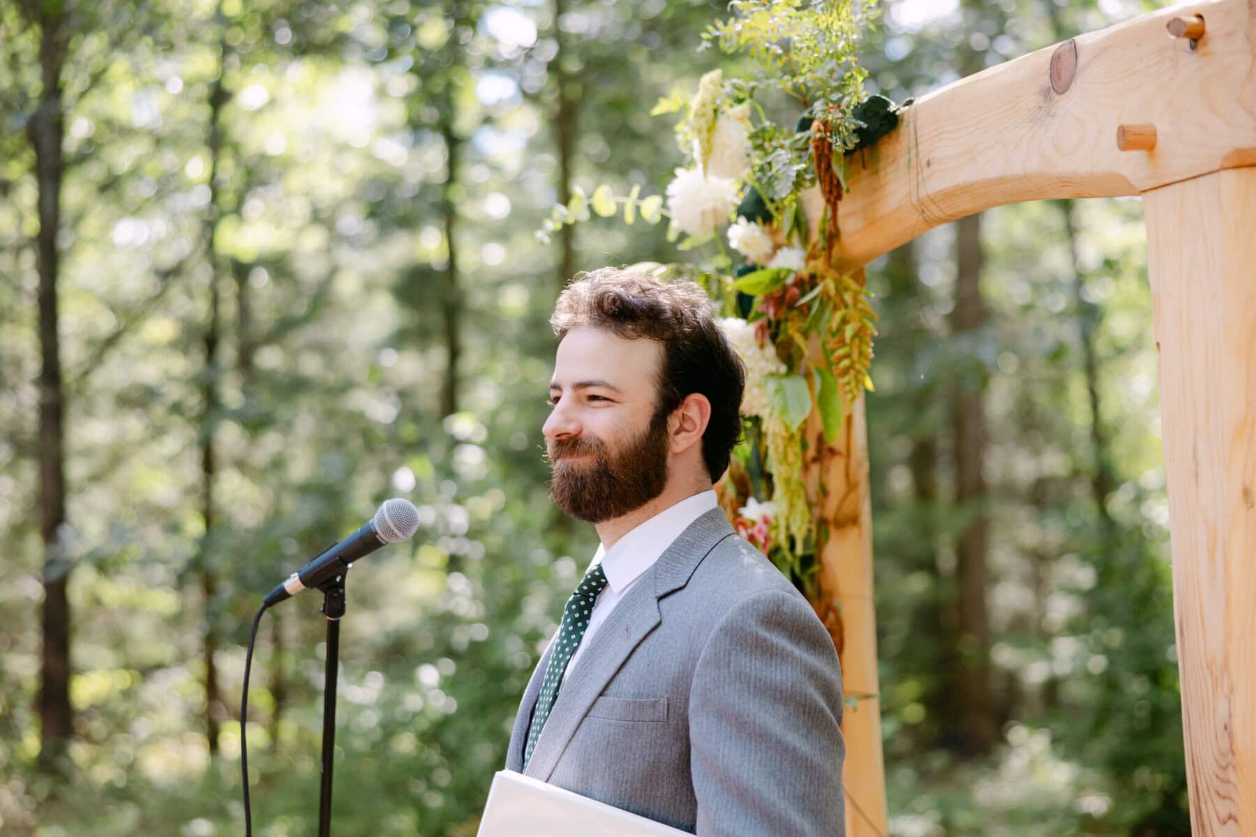 A Worcester Wedding at Cook Pond 24 A man in a gray suit stands at a microphone under a flowered arch, outdoors by Cook Pond at a Worcester Wedding in a forested area.
