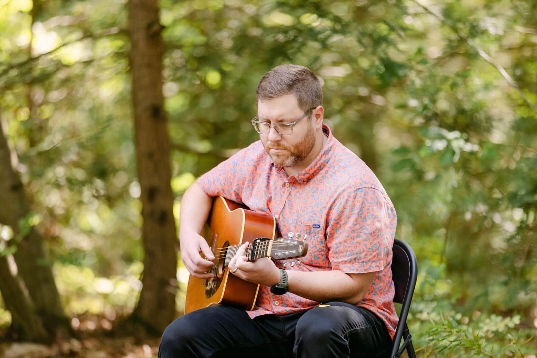A Worcester Wedding at Cook Pond 25 A man with glasses sits on a folding chair at Cook Pond, playing an acoustic guitar in a wooded area.
