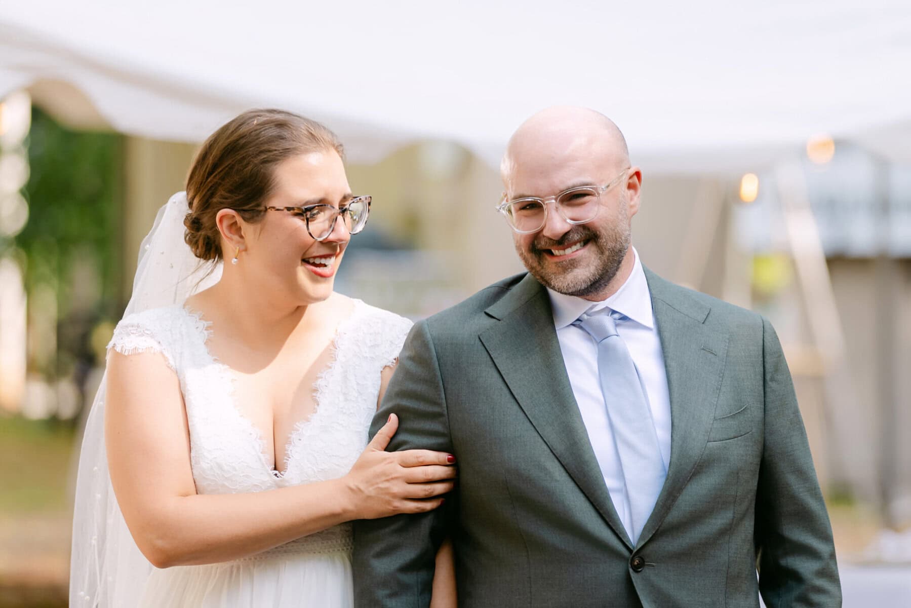A Worcester Wedding at Cook Pond 27 A bride in a white dress and a man in a gray suit smile while walking outdoors under a white canopy at their Worcester Wedding.