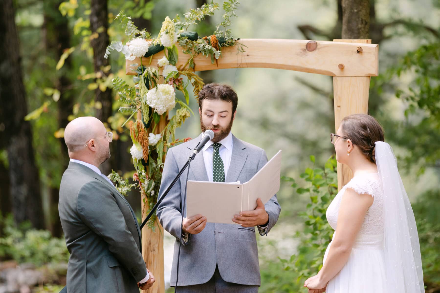 A Worcester Wedding at Cook Pond 29 An officiant reads from a book to a couple during their outdoor Worcester Wedding ceremony under a flower-adorned arch by Cook Pond.