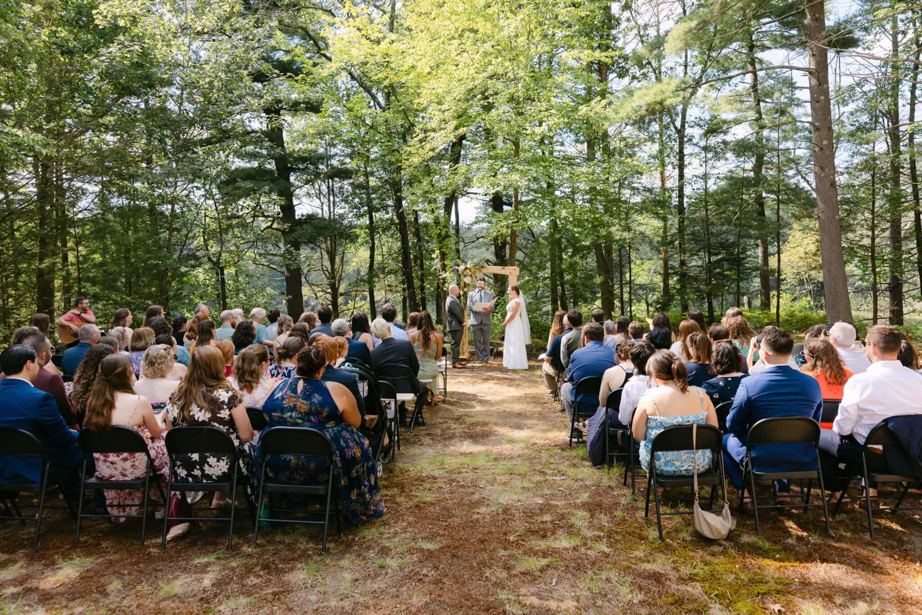 A Worcester Wedding at Cook Pond 30 Outdoor wedding ceremony at Cook Pond, with guests seated in a forest setting as the couple stands with the officiant under a wooden arch.