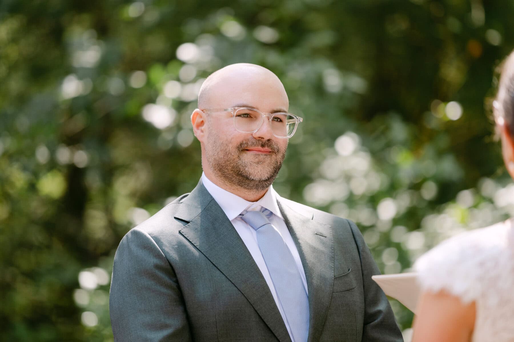 A Worcester Wedding at Cook Pond 31 A man in a suit faces another in white, outdoors at Cook Pond, with lush greenery hinting at a Worcester Wedding in the background.