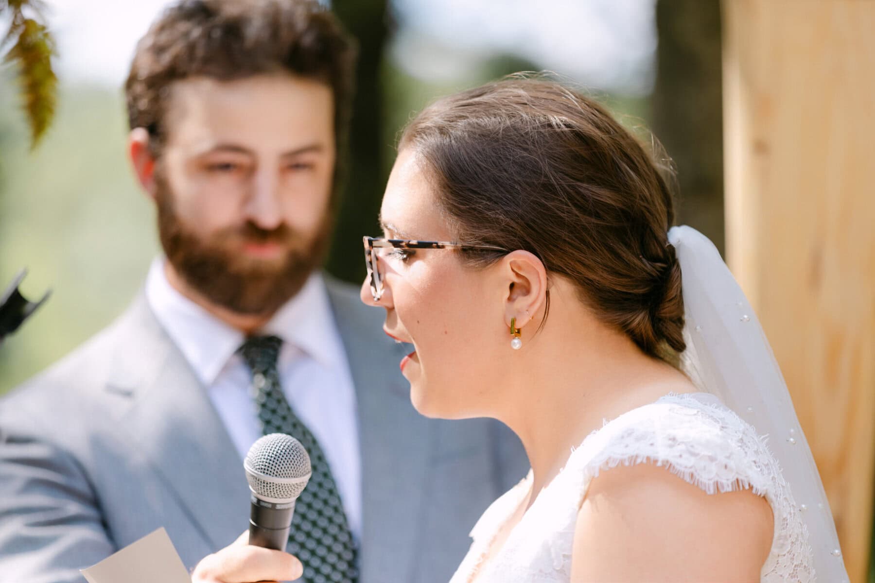 A Worcester Wedding at Cook Pond 34 At a Worcester Wedding by Cook Pond, a woman in a white dress speaks into a microphone as a man in a suit stands beside her holding papers.