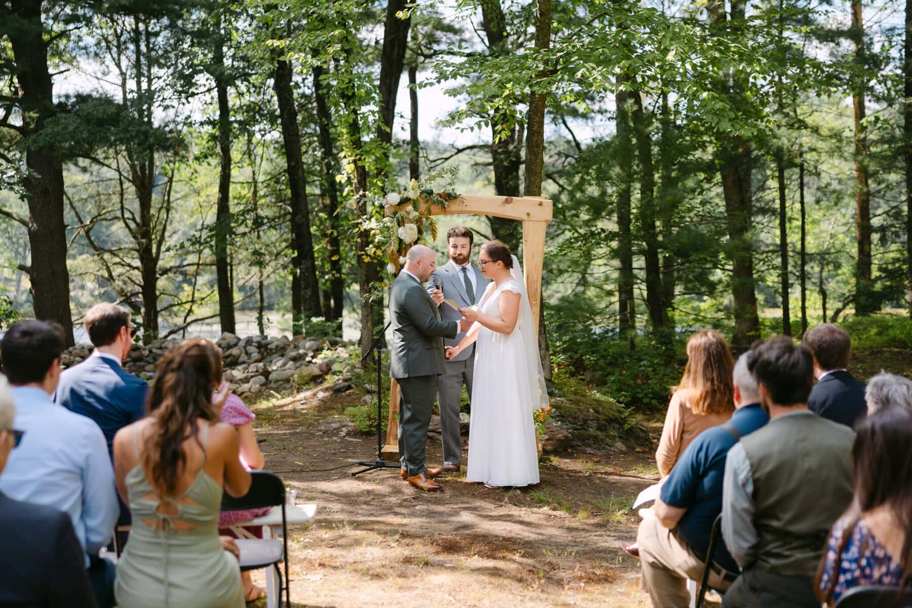 A Worcester Wedding at Cook Pond 36 A couple stands with an officiant under a wooden arch at their Cook Pond wedding, as seated guests observe the outdoor Worcester Wedding ceremony.