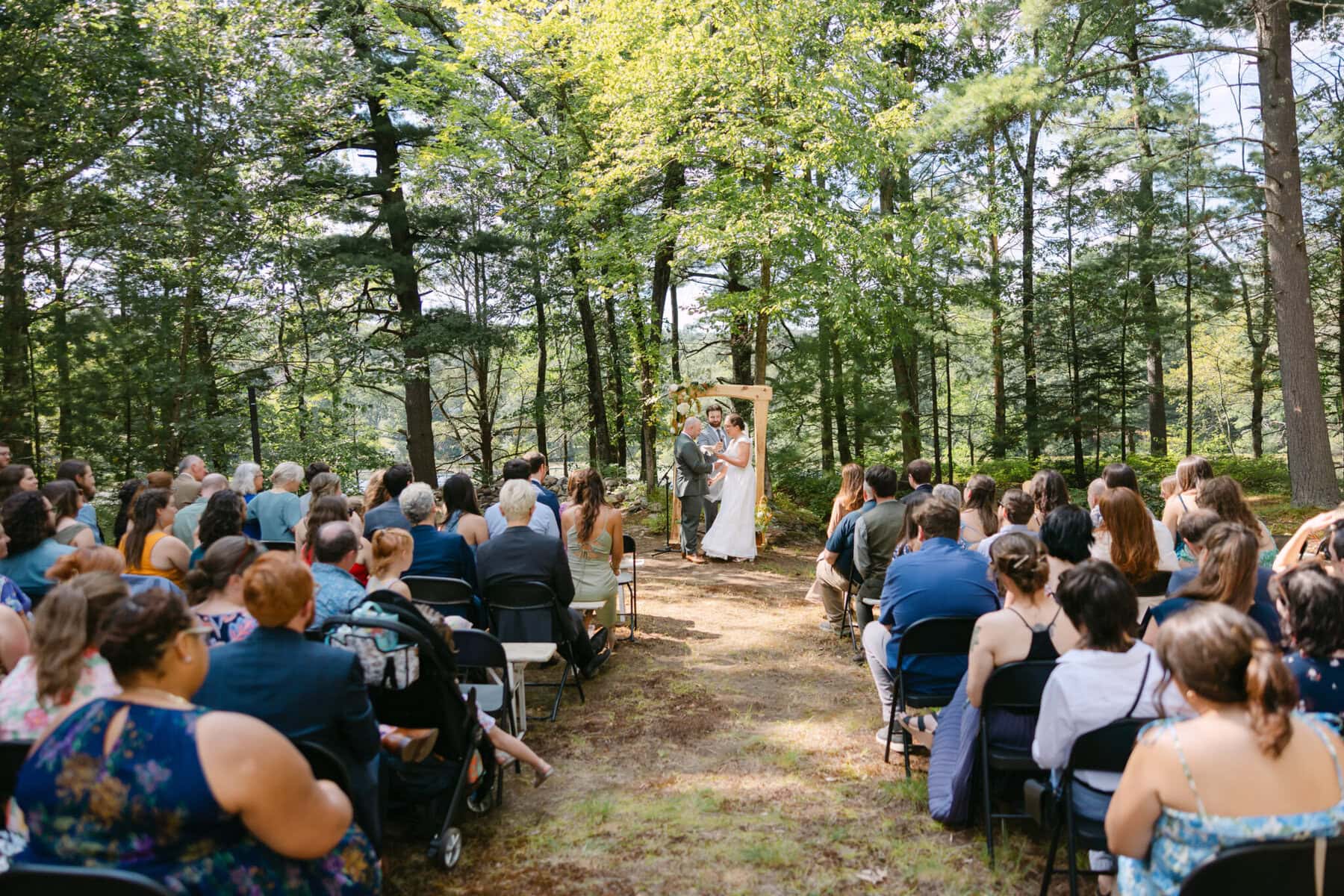 A Worcester Wedding at Cook Pond 37 A couple stands at an outdoor altar by Cook Pond, exchanging vows in front of their Worcester wedding guests among the trees.
