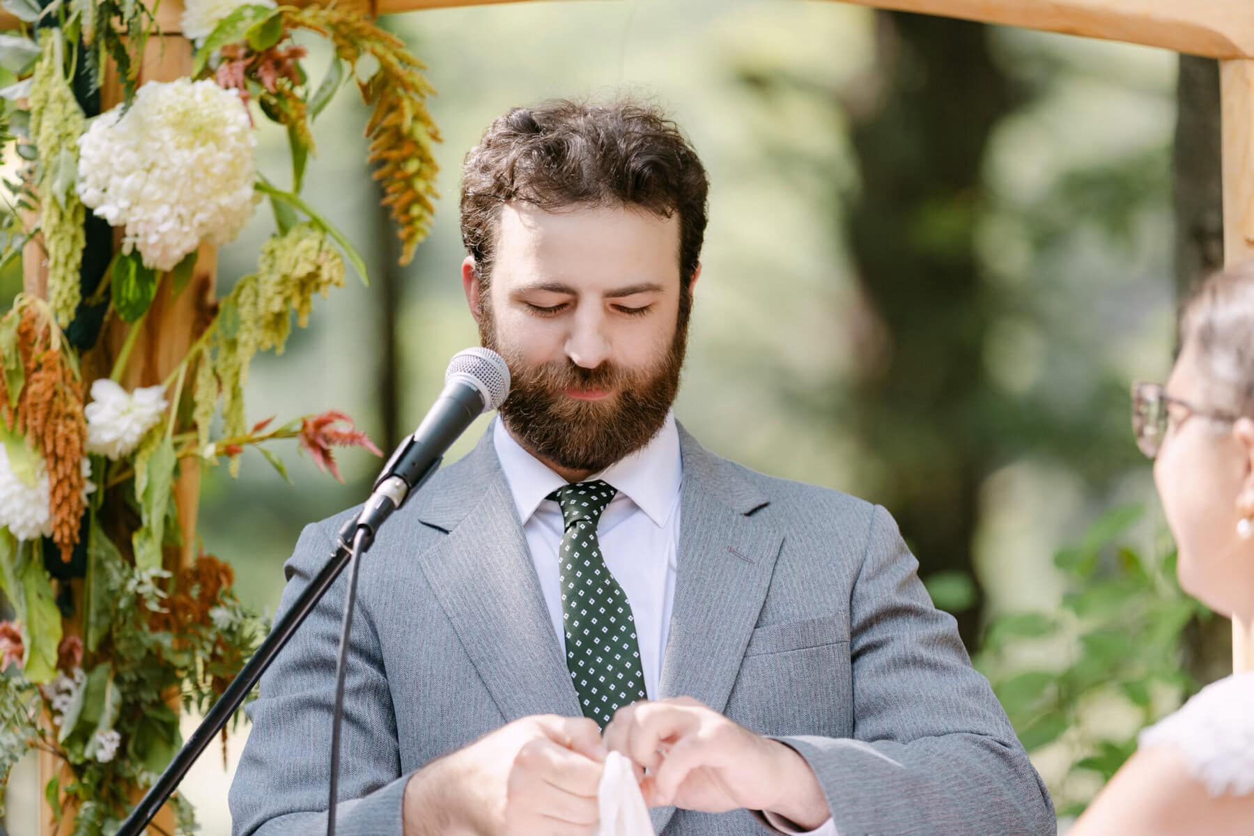 A Worcester Wedding at Cook Pond 39 A man in a gray suit and green polka dot tie stands at a microphone outdoors at a Worcester Wedding, with flowers and greenery behind him.