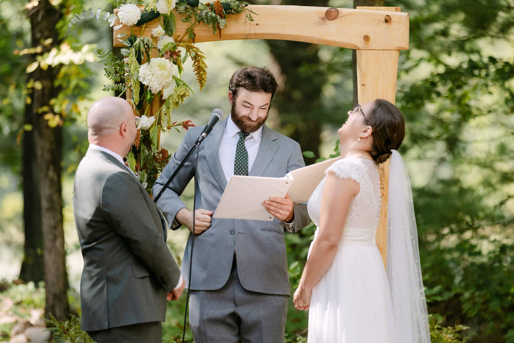 A Worcester Wedding at Cook Pond 41 A couple stands under a wooden arch at Cook Pond as an officiant reads from a script during their Worcester wedding ceremony.