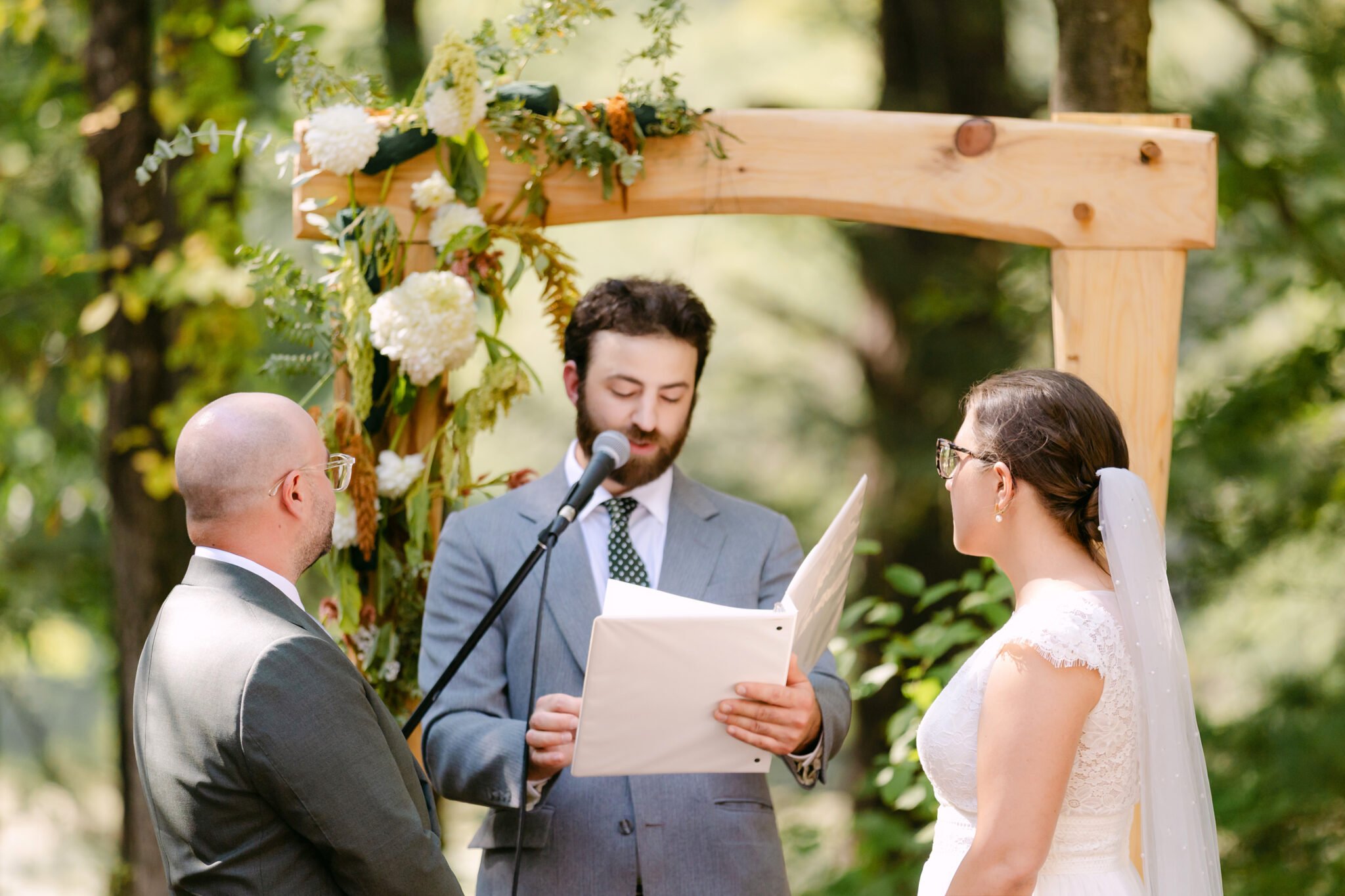 A Worcester Wedding at Cook Pond 42 A man in a suit officiates a Worcester Wedding outdoors, reading from a folder to a bride and groom beneath a wooden arch.