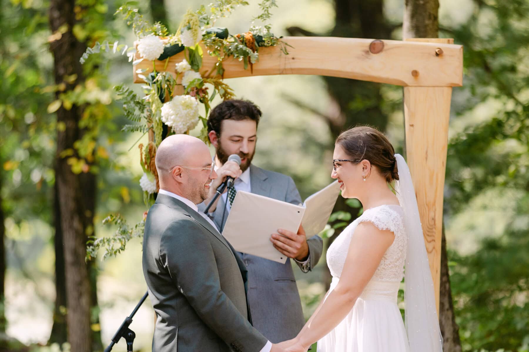 A Worcester Wedding at Cook Pond 43 A couple holds hands and smiles at each other during a Worcester Wedding, with an officiant speaking under a flower-adorned wooden arch.
