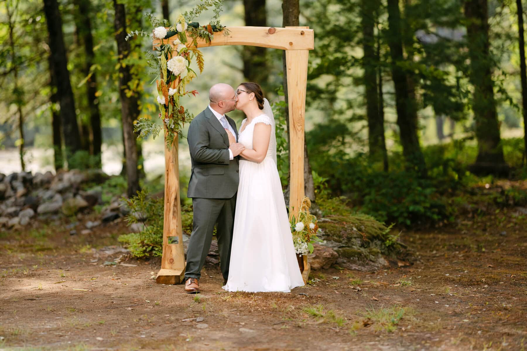 A Worcester Wedding at Cook Pond 51 A bride and groom kiss under a wooden arch with flowers at their enchanting Worcester Wedding in a serene forest setting.