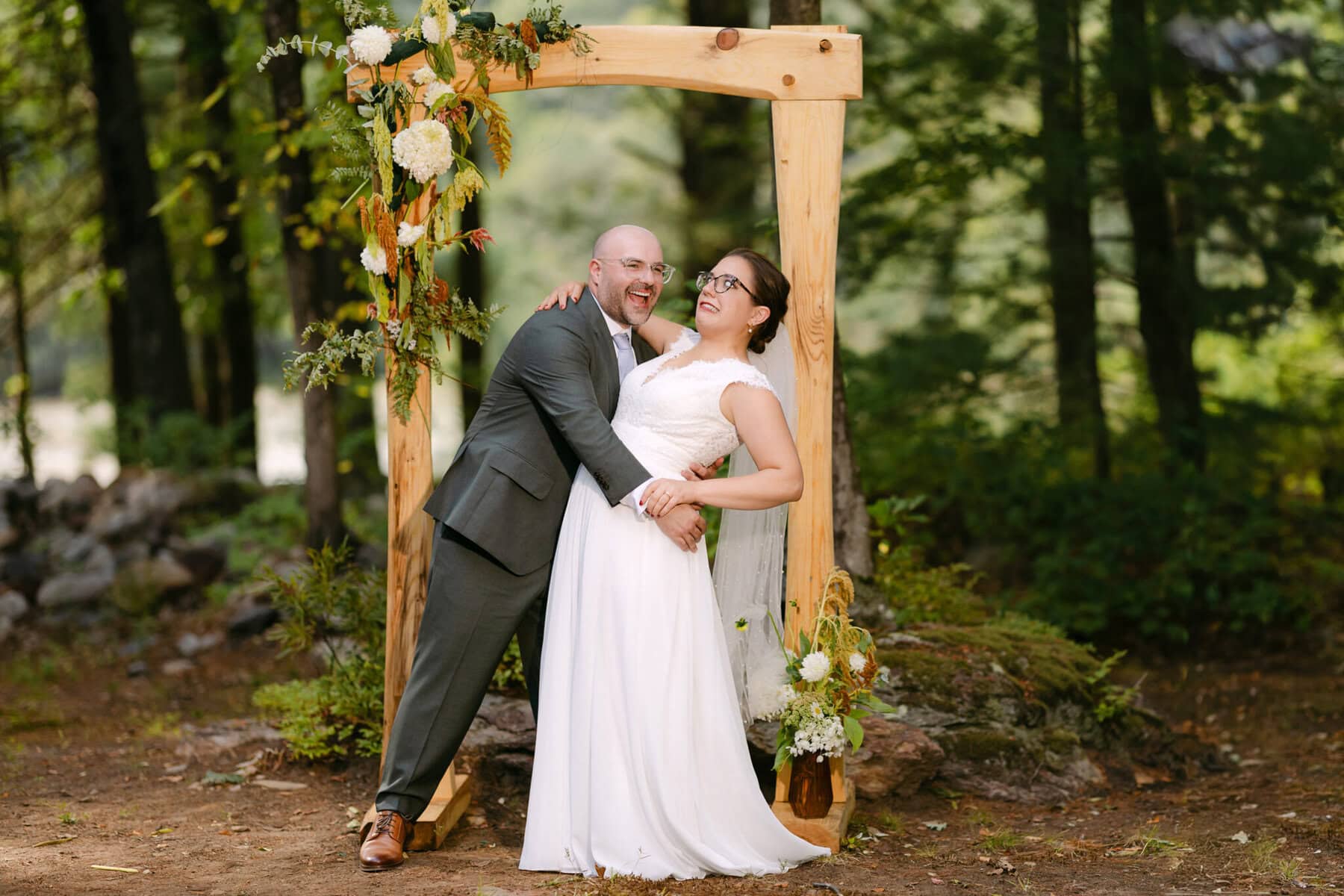 A Worcester Wedding at Cook Pond 52 A bride and groom laugh while posing under a flowered wooden arch in a forest at their enchanting Worcester Wedding.