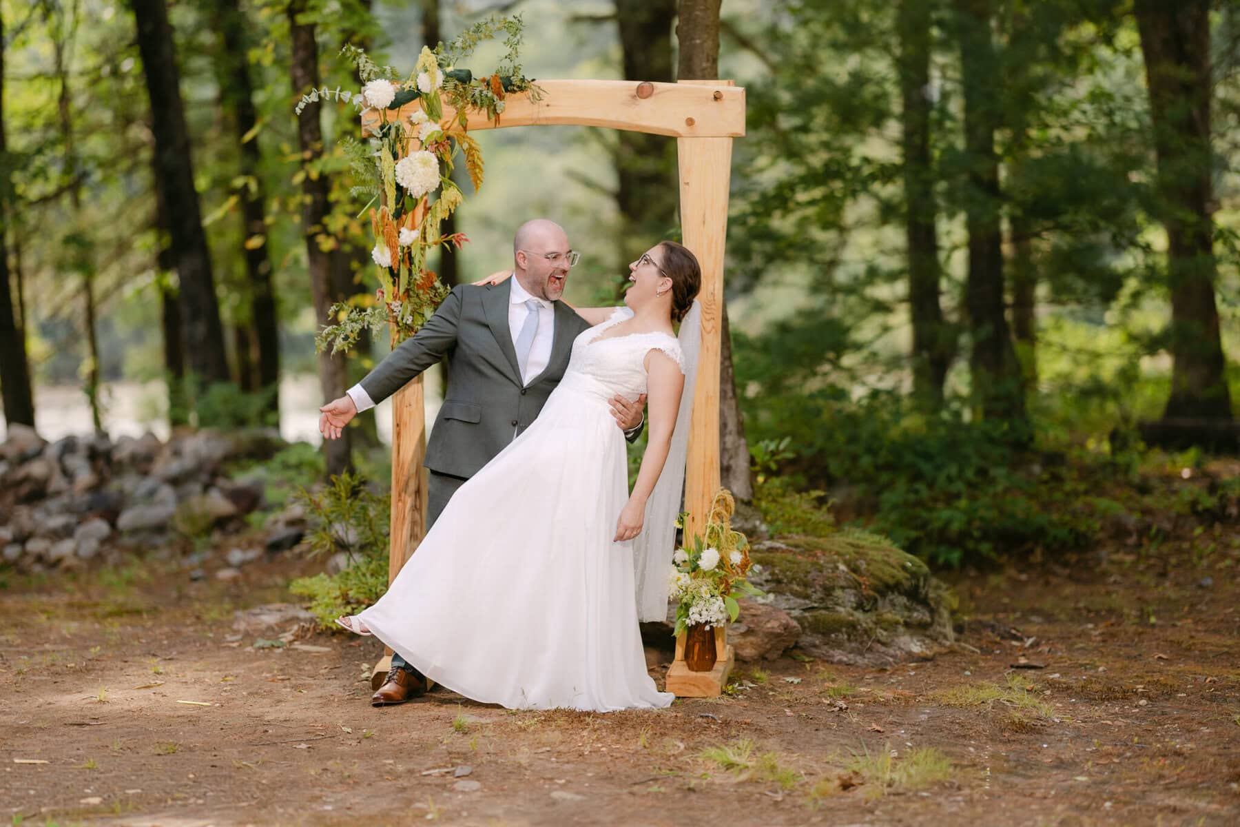 A Worcester Wedding at Cook Pond 53 A bride and groom smile and pose under a flower-adorned wooden arch in a wooded outdoor setting at their Worcester Wedding.