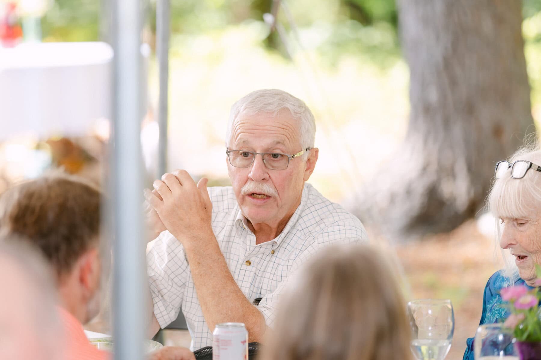 A Worcester Wedding at Cook Pond 58 An older man with glasses speaks at a Cook Pond gathering, surrounded by guests seated at a festive table.