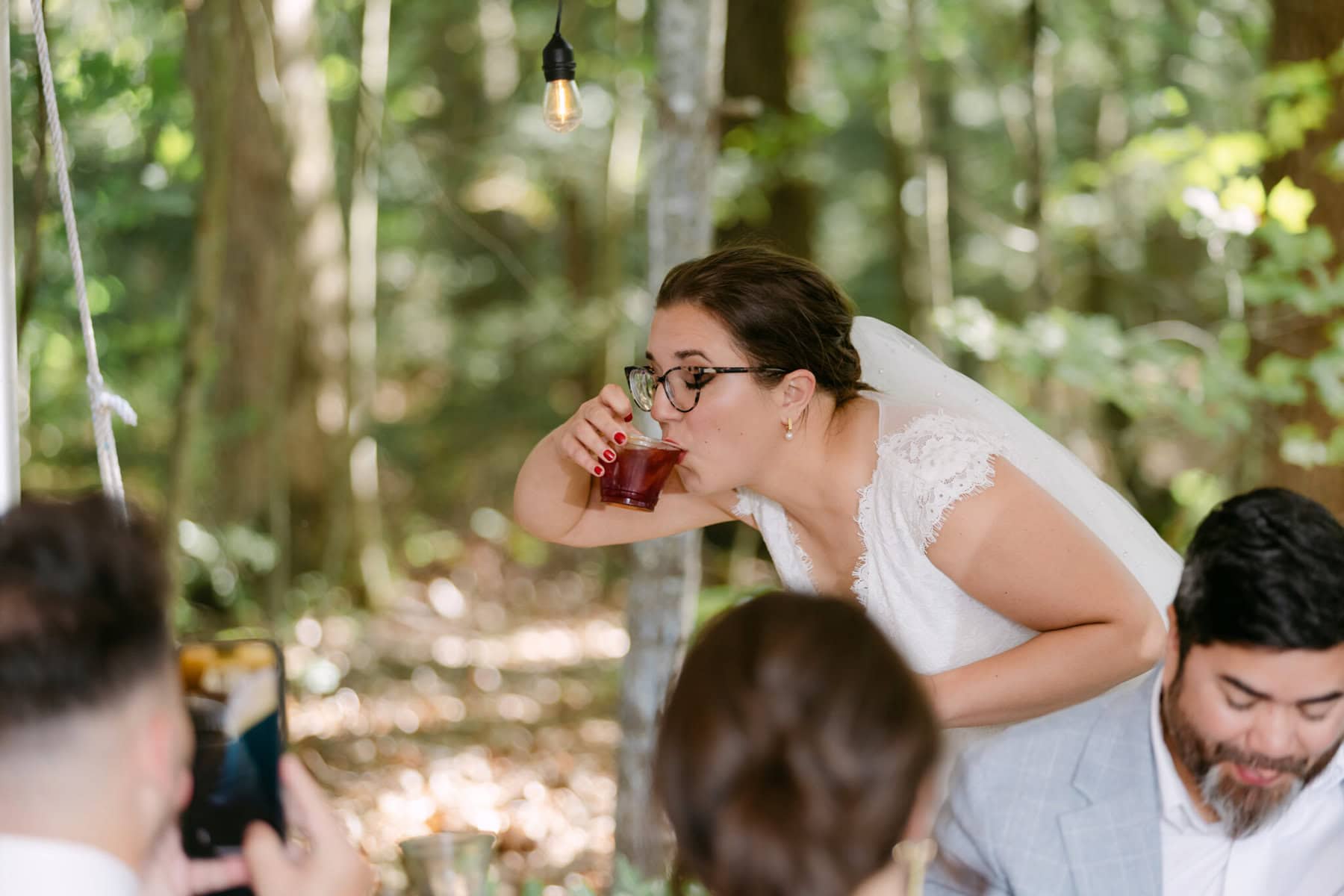 A Worcester Wedding at Cook Pond 59 A bride in a white dress and glasses drinks from a glass outdoors at a Worcester Wedding, surrounded by trees and guests.