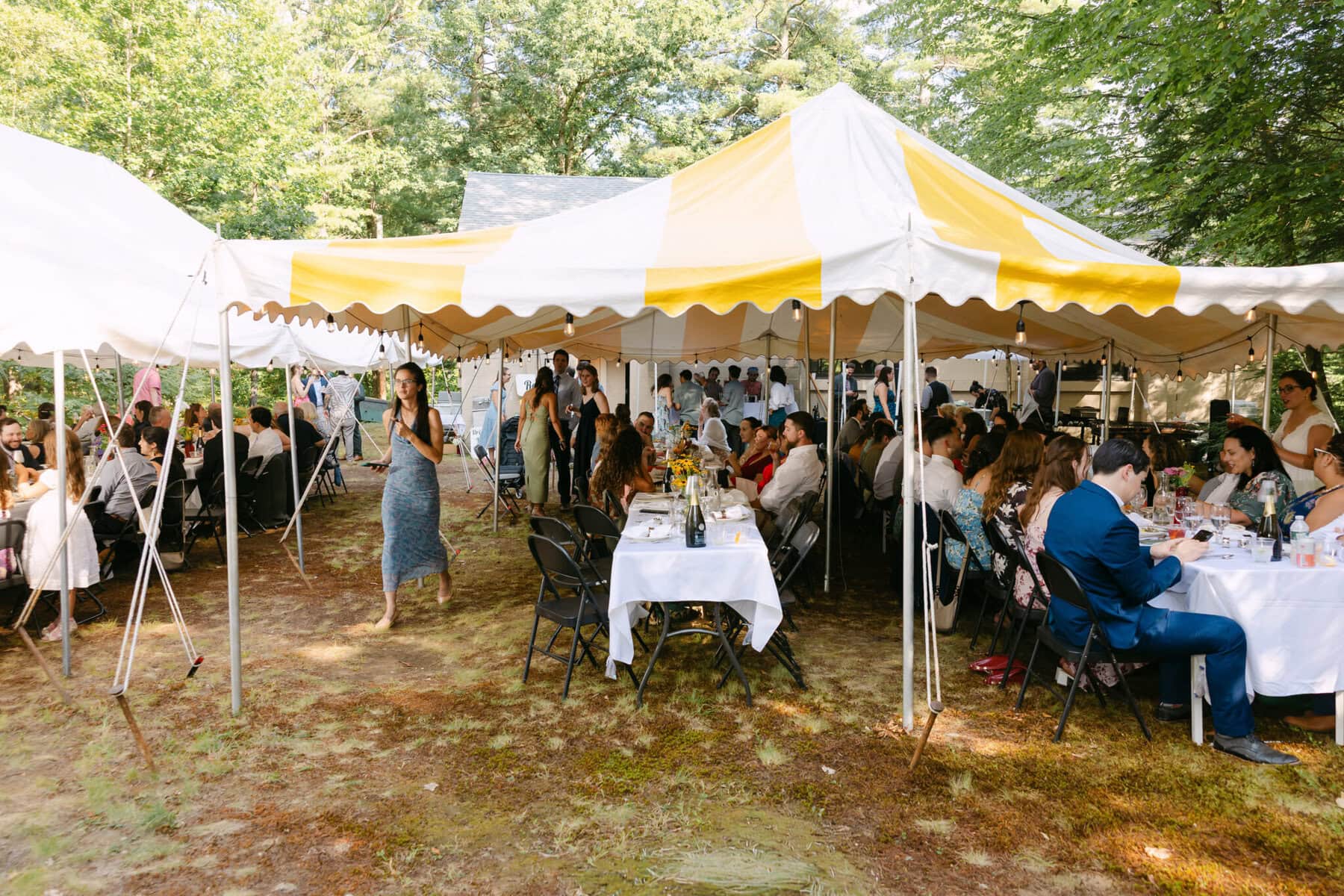 A Worcester Wedding at Cook Pond 60 People sit and eat at round tables under a large tent at a sunny outdoor Cook Pond event.