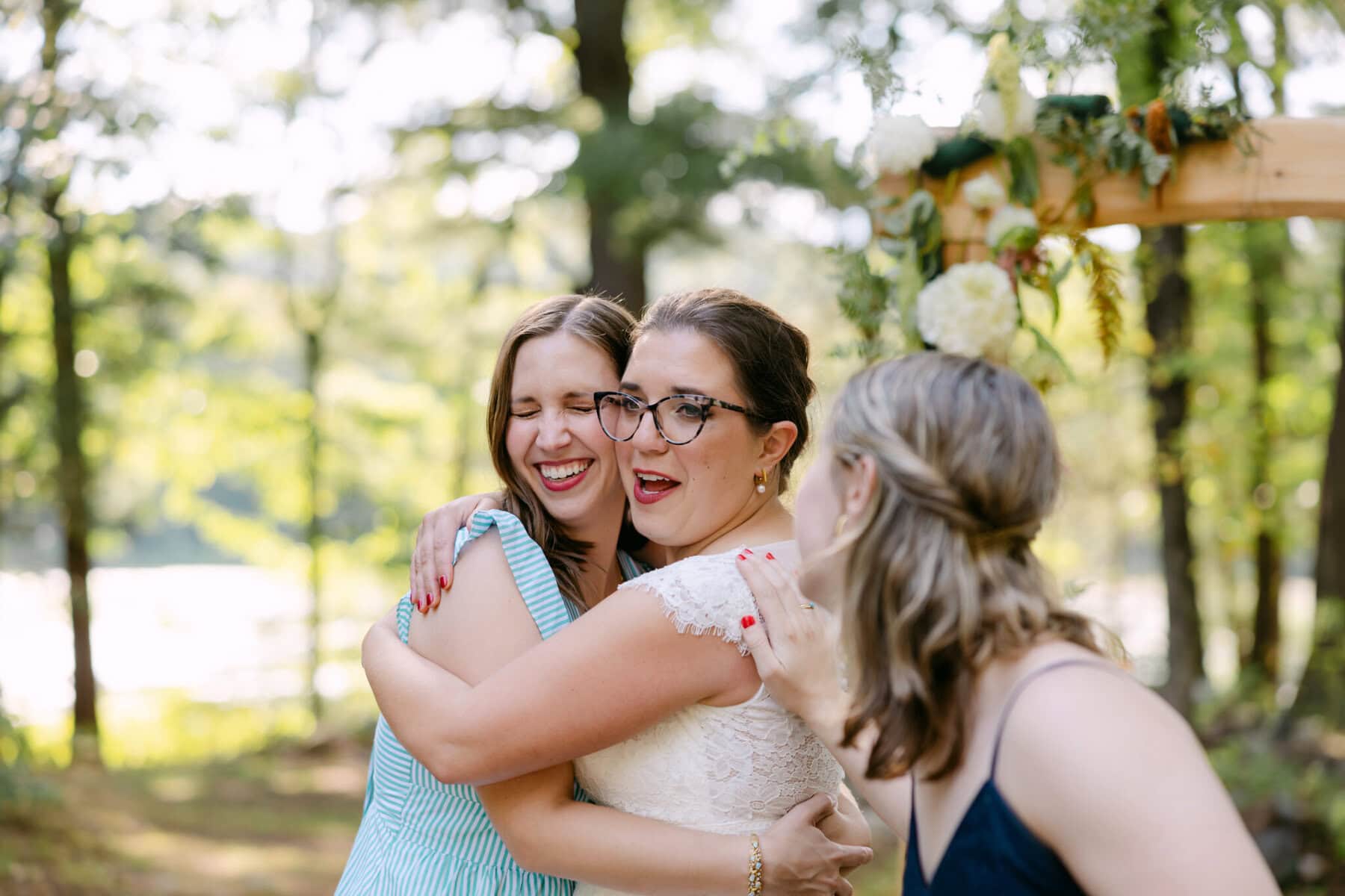 A Worcester Wedding at Cook Pond 62 Three women hug and smile outdoors near a flower-adorned arch at a Worcester Wedding, with trees and sunlight in the background.
