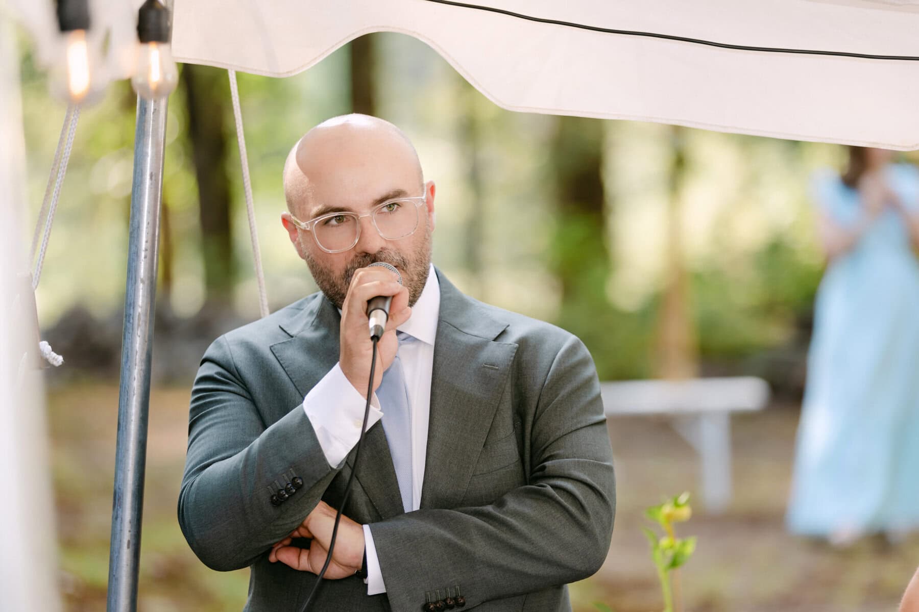 A Worcester Wedding at Cook Pond 63 A man in a suit and glasses speaks into a microphone at a Worcester Wedding under a white canopy, with trees and guests in the background.