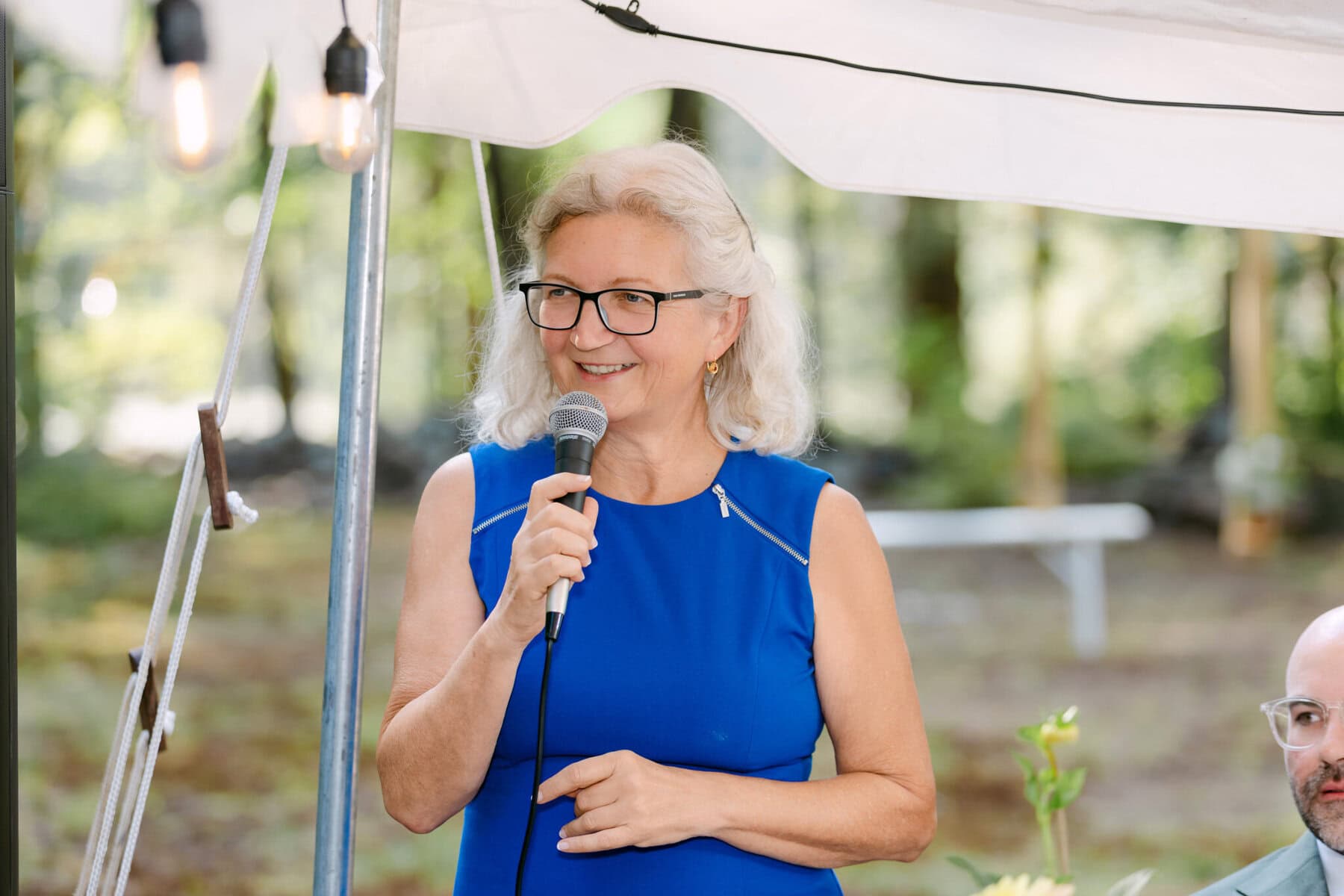 A Worcester Wedding at Cook Pond 64 A woman with gray hair and glasses speaks into a microphone under a white tent at a Worcester Wedding, wearing a blue sleeveless dress.