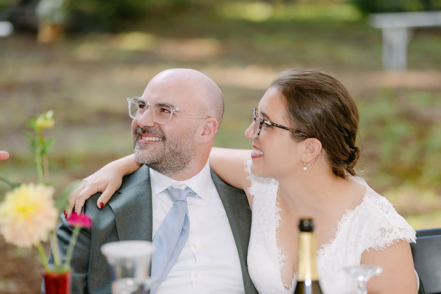 A Worcester Wedding at Cook Pond 65 A man in a suit and a woman in a white dress share a smile at their Worcester Wedding, sitting close together outdoors by Cook Pond.