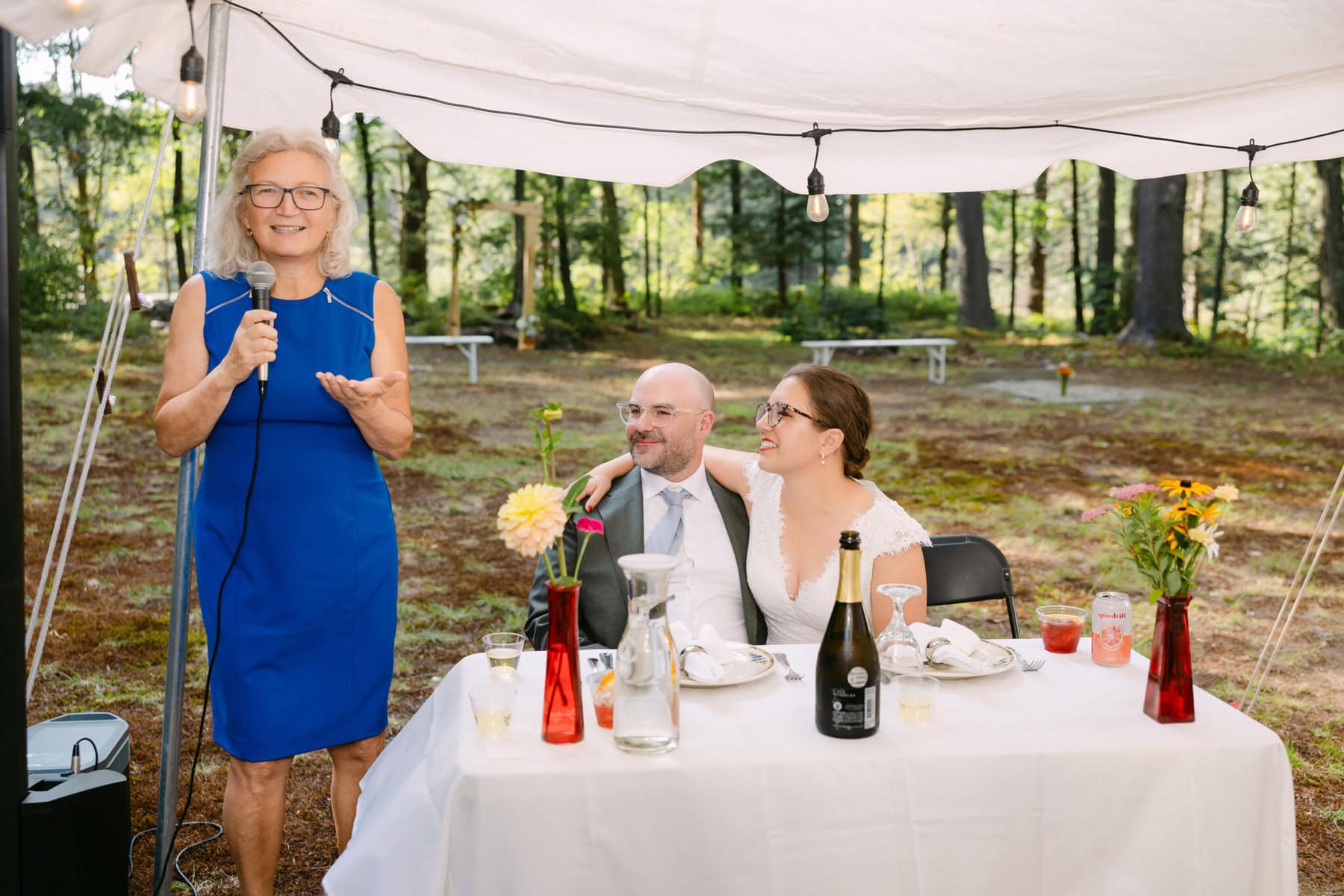 A Worcester Wedding at Cook Pond 66 At a Worcester Wedding by Cook Pond, a woman in a blue dress speaks into a microphone beside the happy couple at their outdoor reception table.