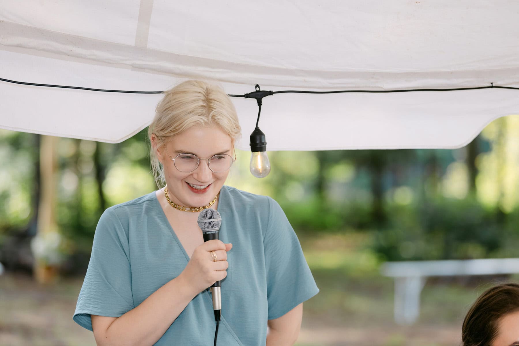 A Worcester Wedding at Cook Pond 67 A person with blond hair and glasses speaks into a microphone at a Worcester Wedding under a white canopy, greenery in the background.