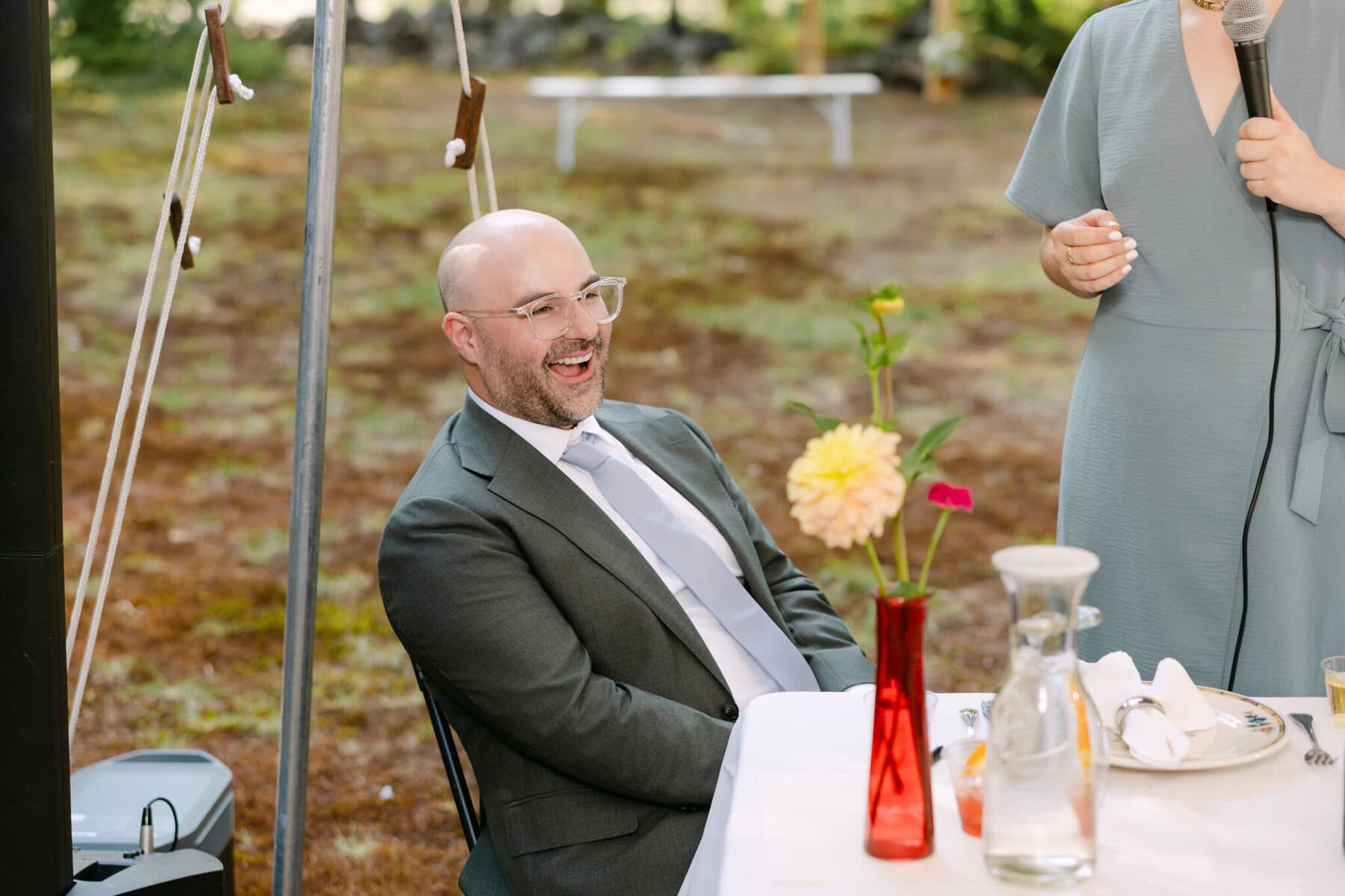 A Worcester Wedding at Cook Pond 68 A man in a suit sits at a table outdoors by Cook Pond, smiling, while a person next to him speaks into a microphone at a Worcester Wedding.