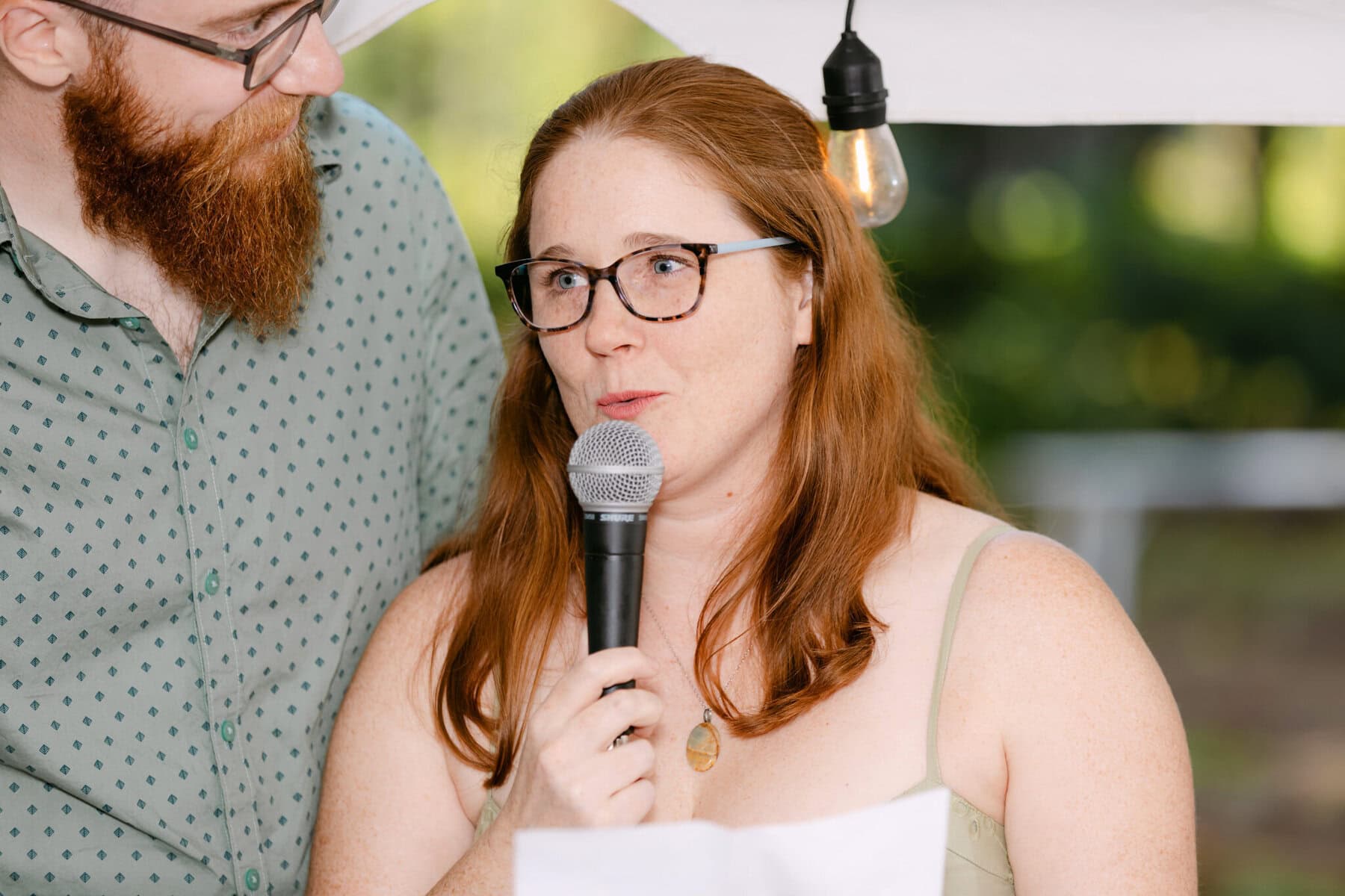 A Worcester Wedding at Cook Pond 69 A woman with red hair and glasses speaks into a microphone beside a bearded man at an outdoor Worcester Wedding.