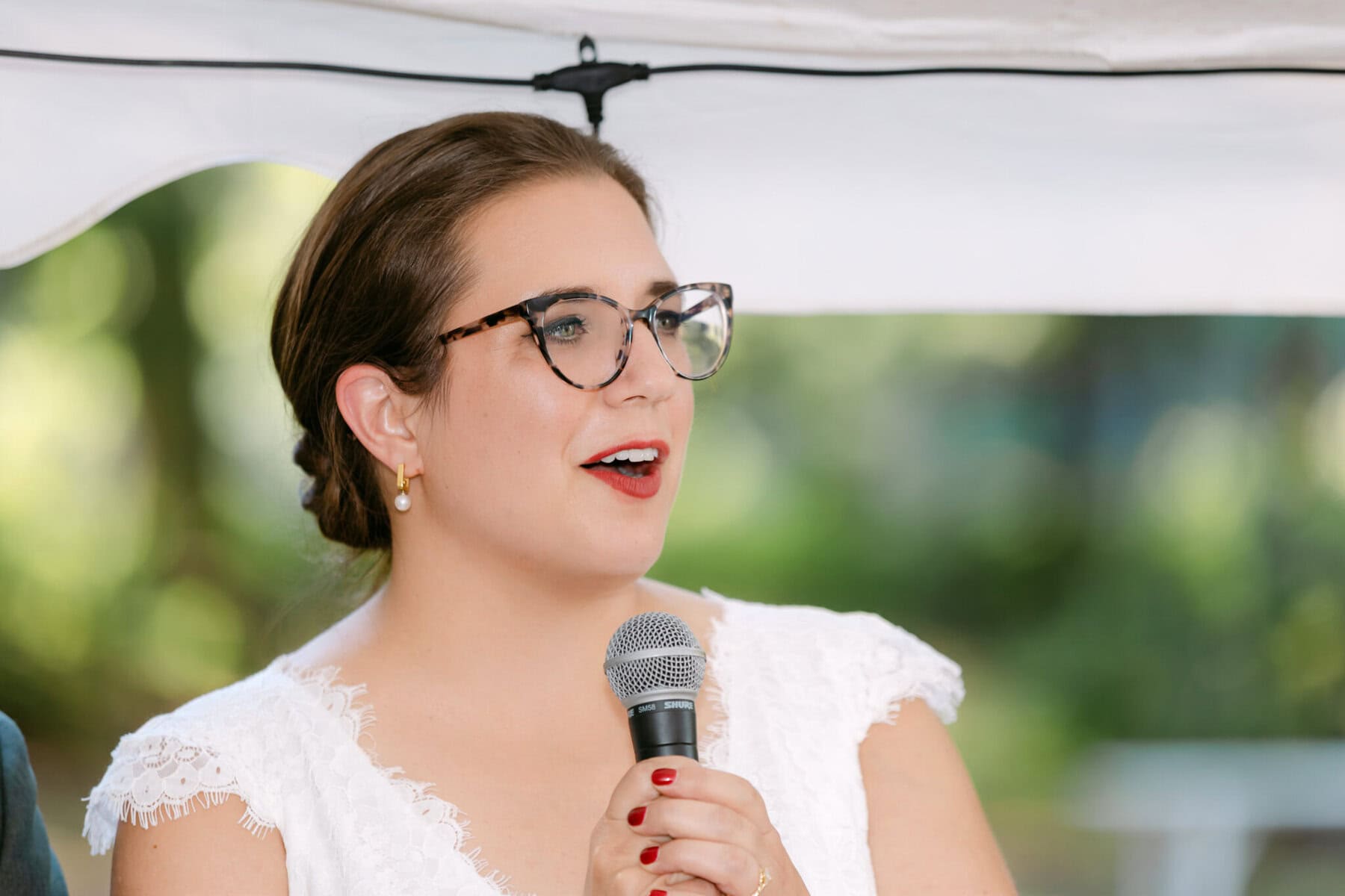 A Worcester Wedding at Cook Pond 70 A woman with glasses in a white dress speaks into a microphone at a beautiful Worcester Wedding under a white canopy.