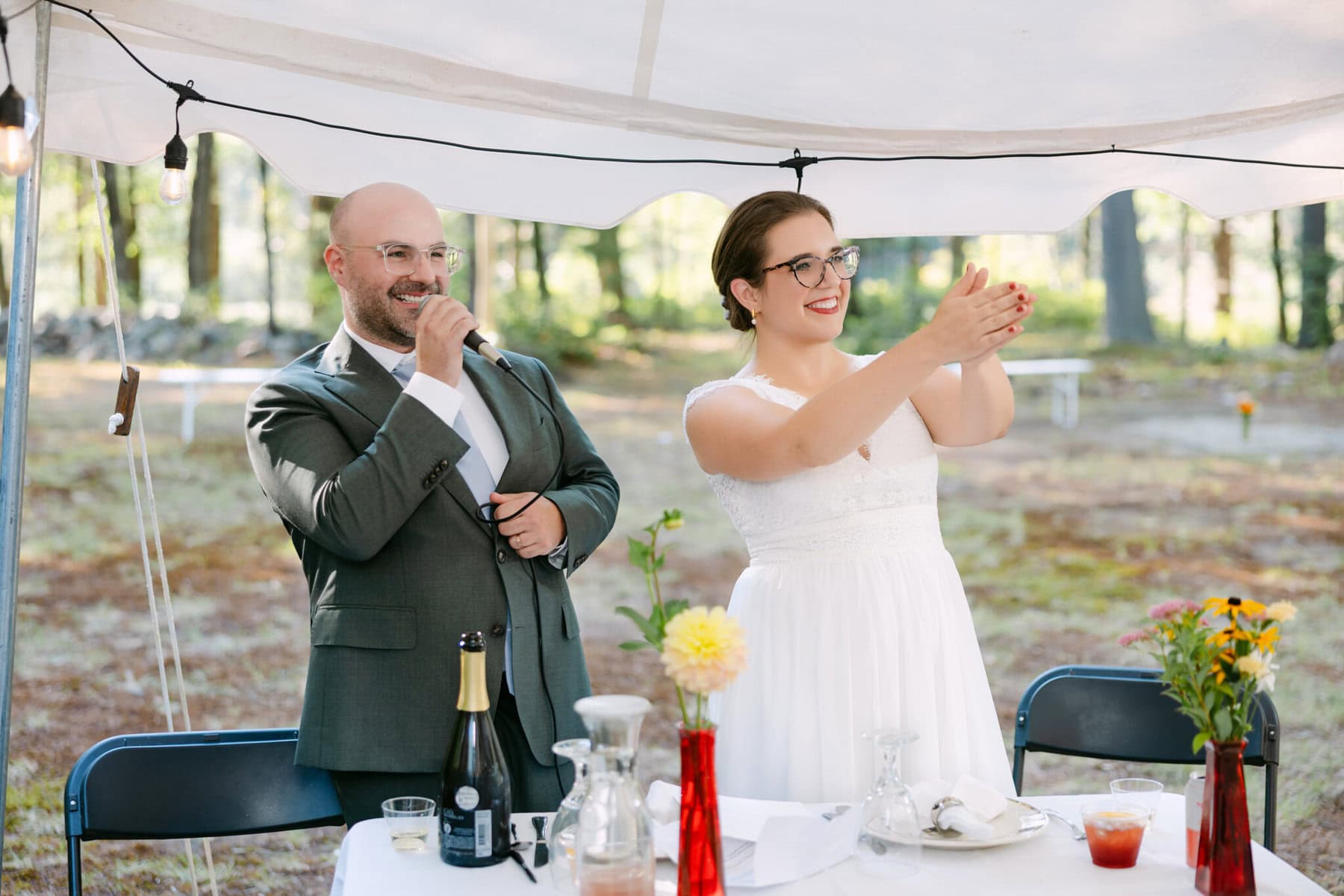 A Worcester Wedding at Cook Pond 71 A man and woman stand under a tent at a decorated table at a Worcester Wedding; she claps as he smiles, holding a microphone.
