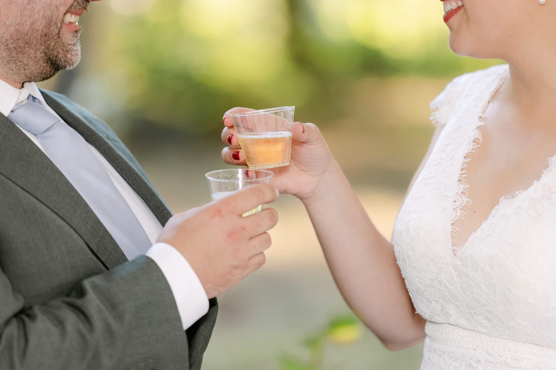 A Worcester Wedding at Cook Pond 73 A man in a suit and a woman in a white lace dress share smiles and plastic cups at a Worcester Wedding by Cook Pond.