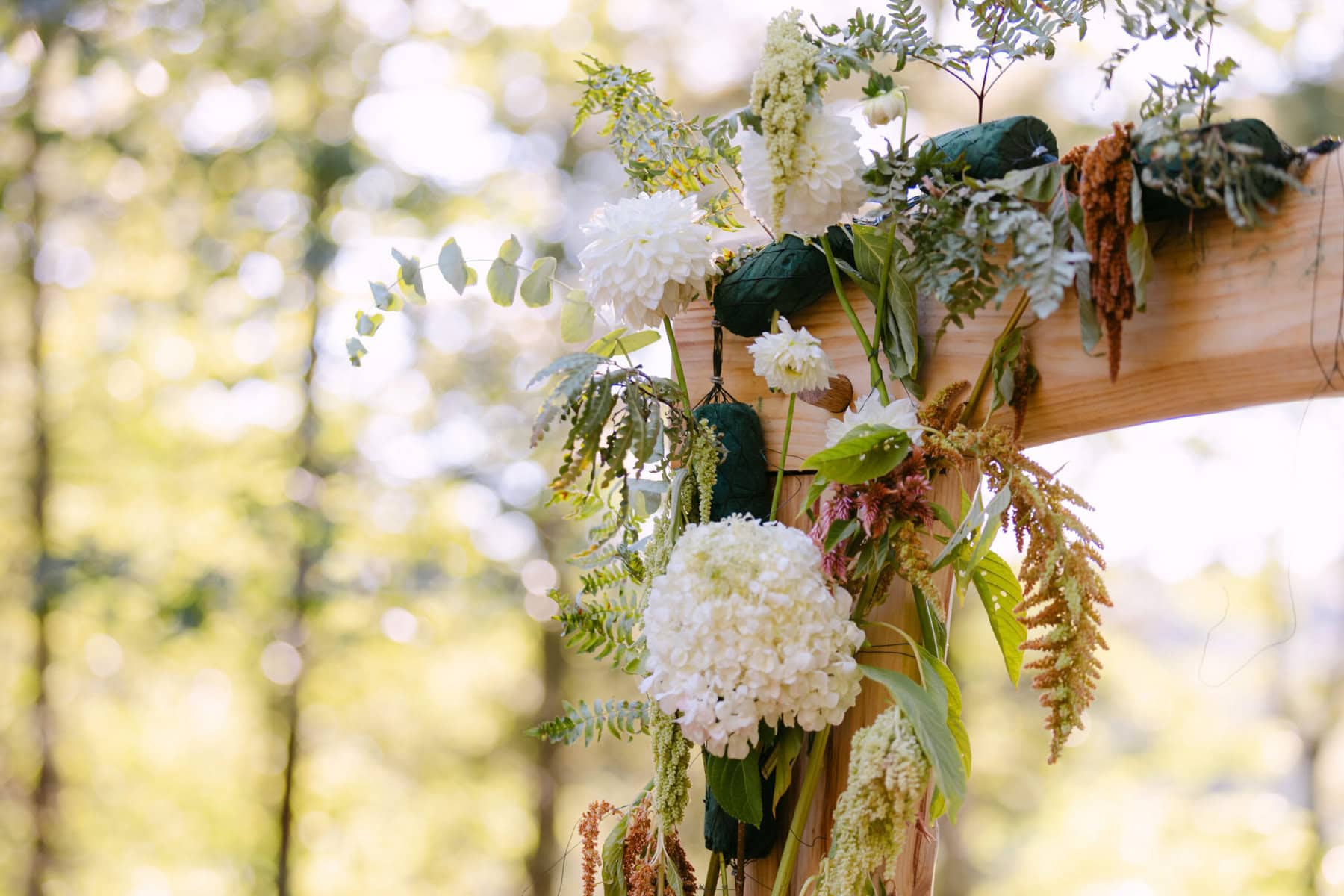 A Worcester Wedding at Cook Pond 74 A wooden arch adorned with white flowers and ferns, perfect for a Worcester Wedding, set outdoors with blurred trees in the background.