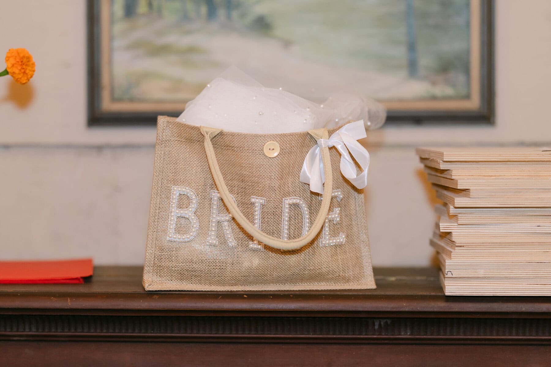 A Worcester Wedding at Cook Pond 75 A beige "BRIDE" bag with a white bow rests on a wooden table at a Worcester Wedding, beside books and an orange flower.
