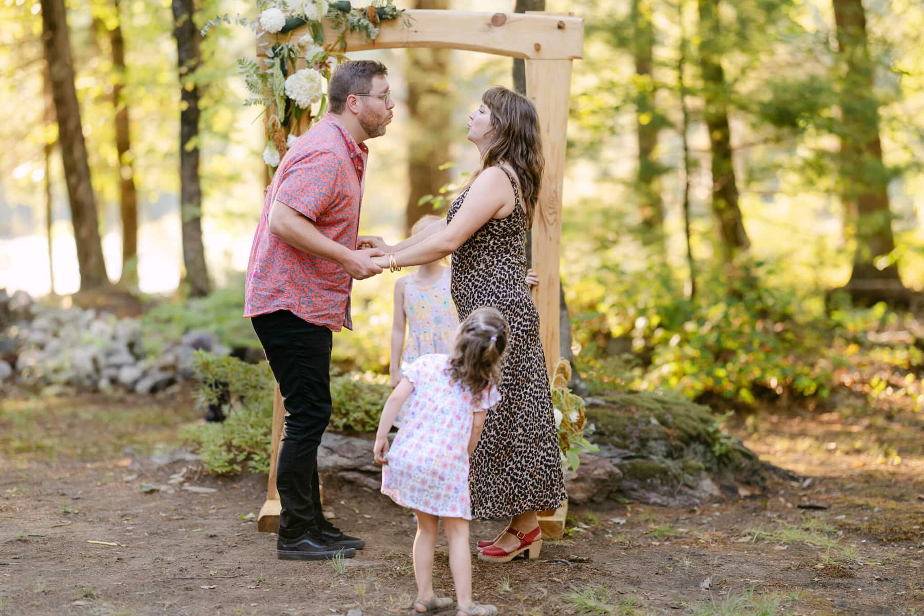 A Worcester Wedding at Cook Pond 76 A couple holds hands under a wooden arch at their Worcester Wedding, with two young girls in front of them near Cook Pond.