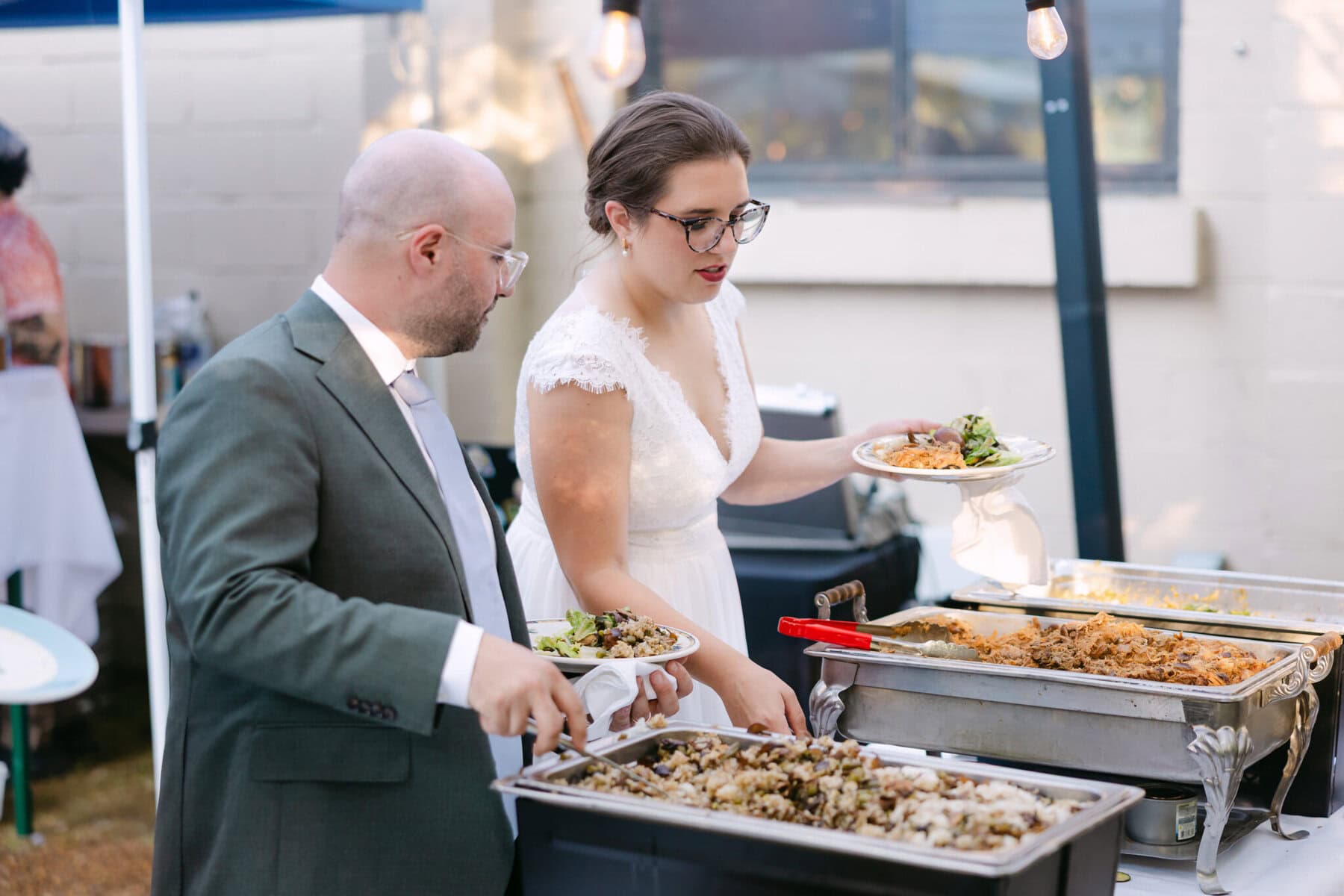 A Worcester Wedding at Cook Pond 77 A man in a suit and a woman in a white dress serve themselves food at an outdoor Worcester Wedding buffet table.