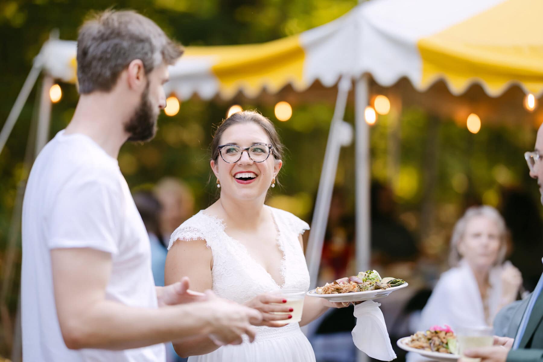 A Worcester Wedding at Cook Pond 78 A woman in a white dress smiles with a plate of food while chatting at a Worcester Wedding under a yellow-and-white tent.